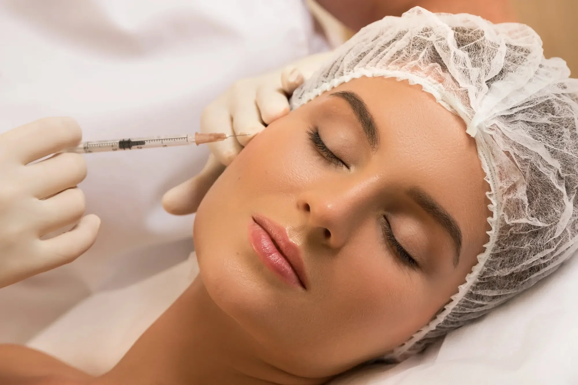 A woman is lying down with her eyes closed, receiving an injection in her face, possibly a cosmetic procedure. She is wearing a protective hair cover and medical gloves, with a professional administering the injection.