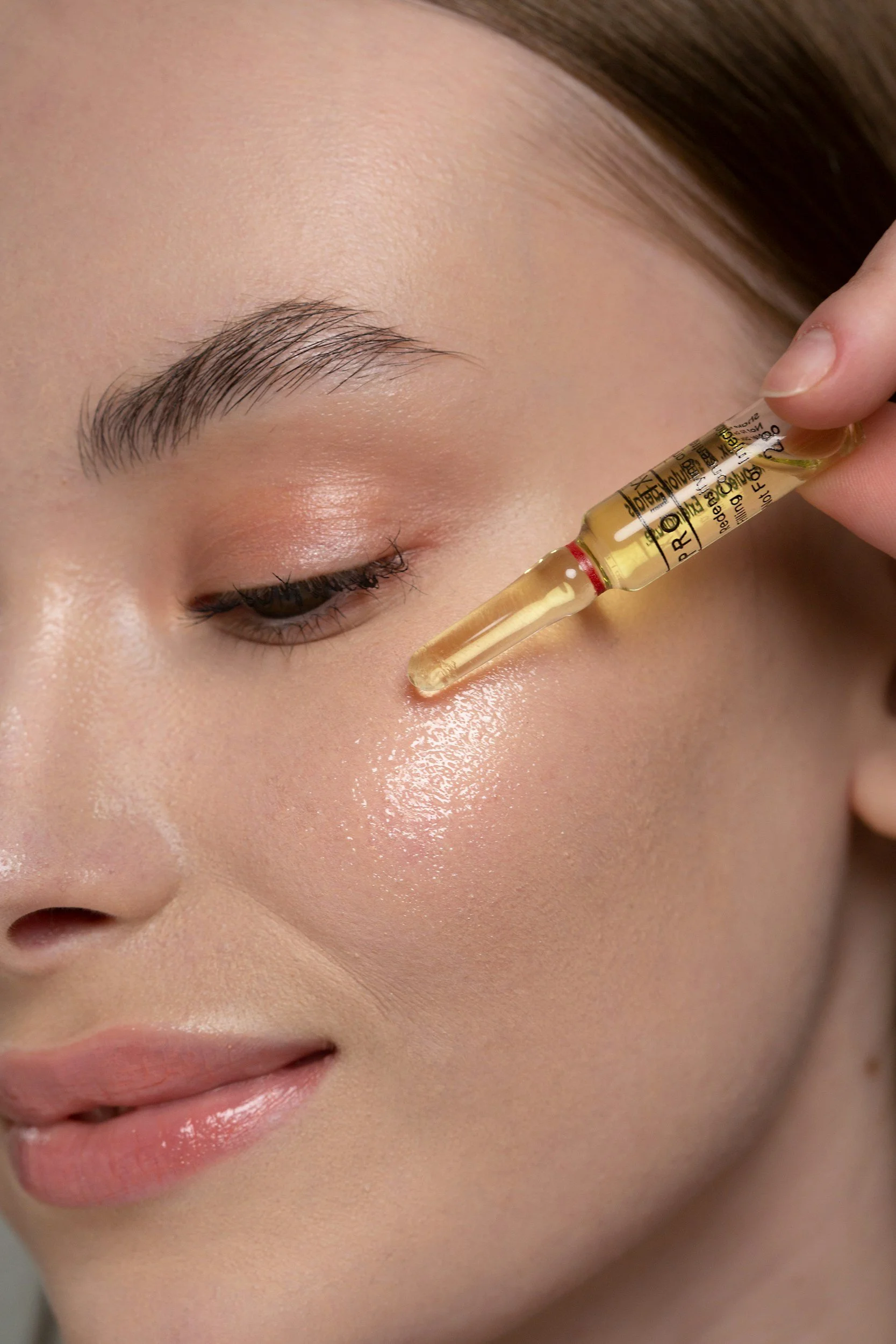 Close-up of a person's face with closed eyes, as a dropper applies a serum or oil to their cheek.