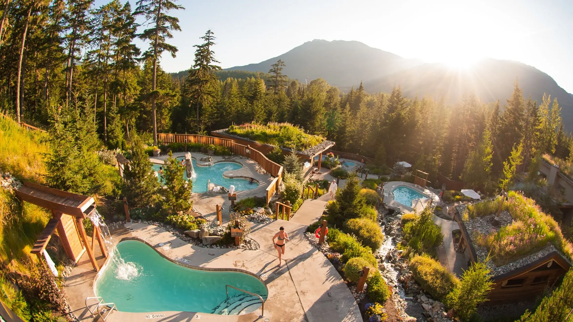 A scenic outdoor hot spring resort with two swimming pools, surrounded by lush trees and shrubs, with mountains in the background and the sun shining brightly in the sky.