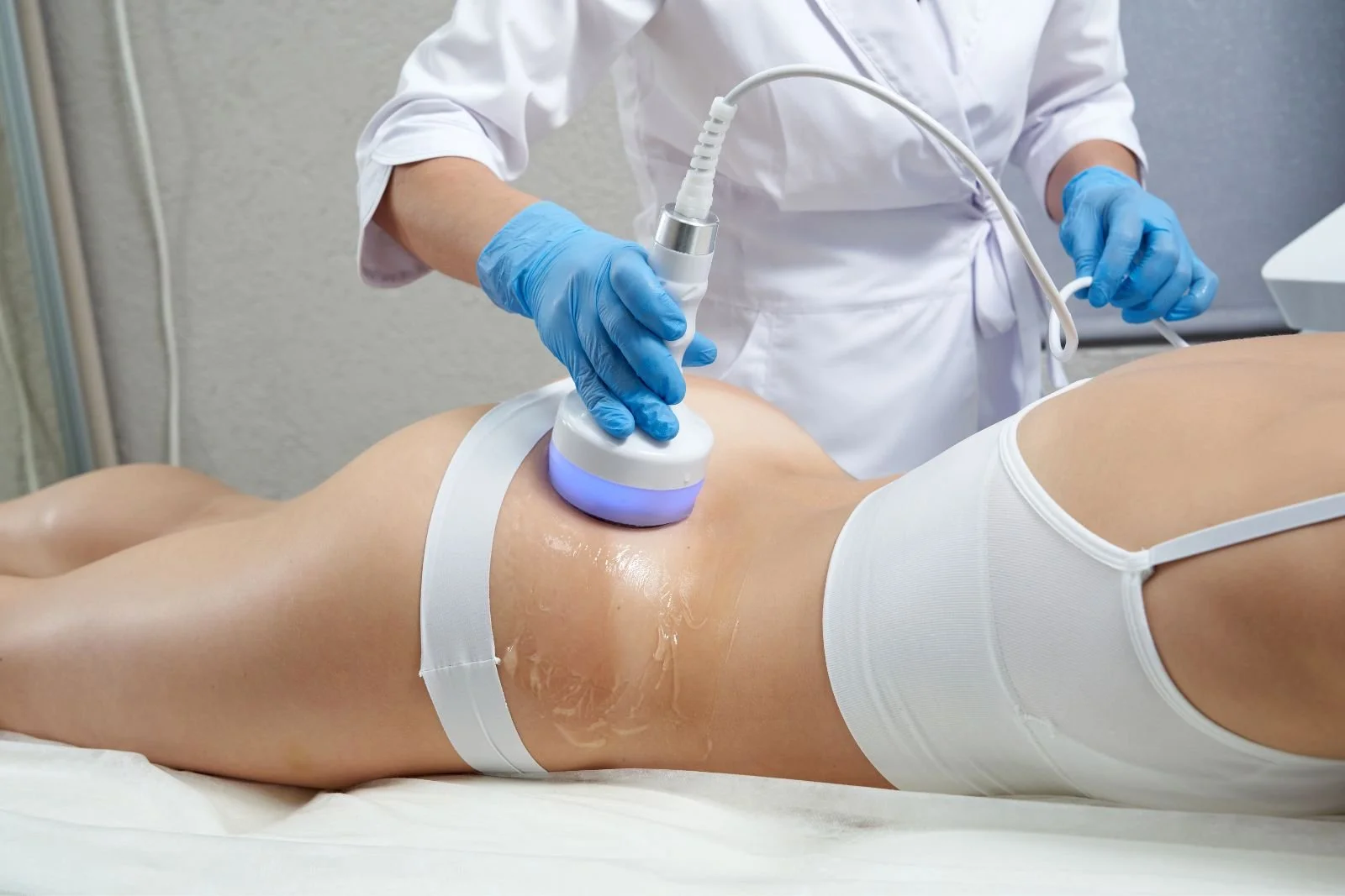 A person lying on a medical table receiving a treatment with a device held by a healthcare professional wearing blue gloves, white coat, and white underwear, in a clinical setting.