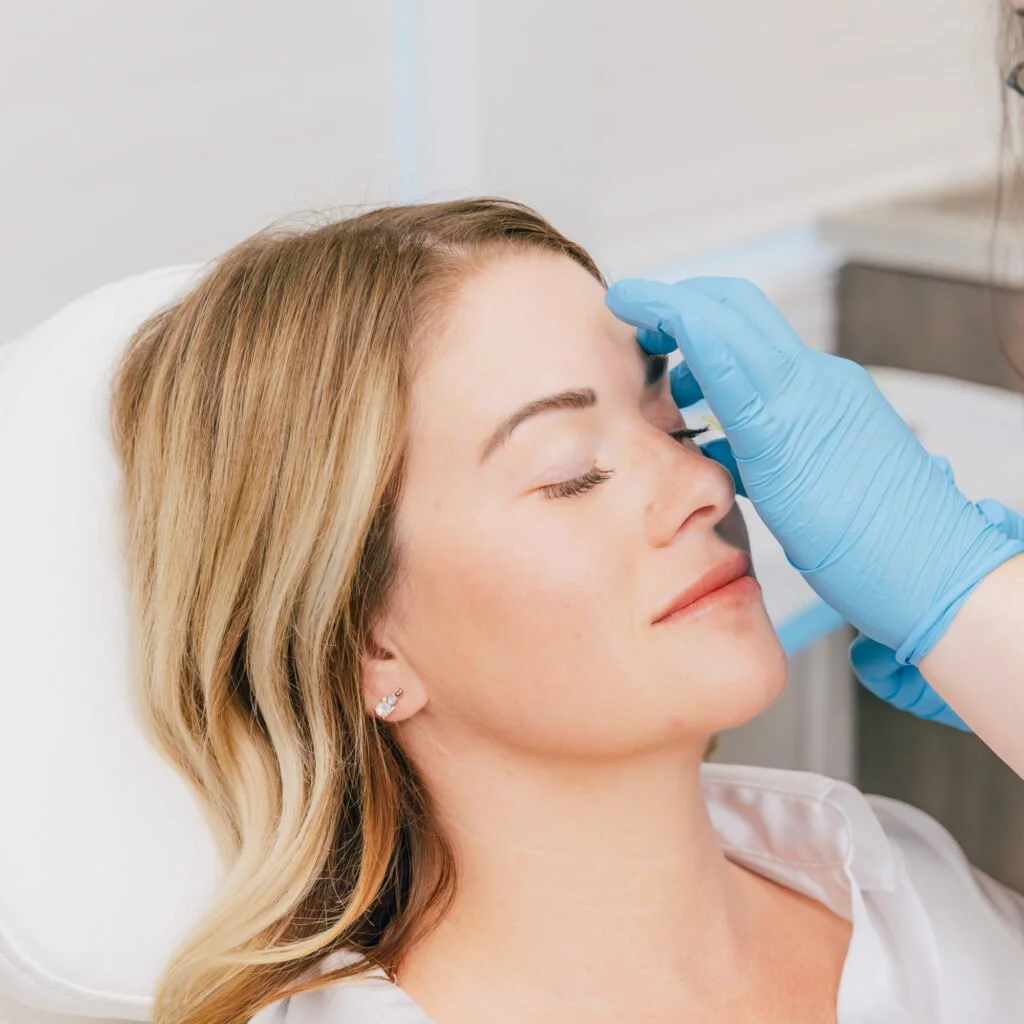 A woman receiving a cosmetic treatment on her forehead from a professional wearing blue gloves.