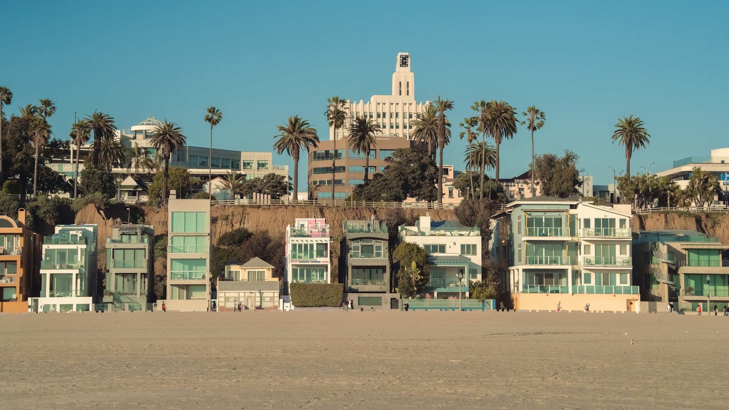 Beachside row of modern multi-story houses with large glass windows, set against a dirt hill, with palm trees and city buildings in the background under a clear blue sky.