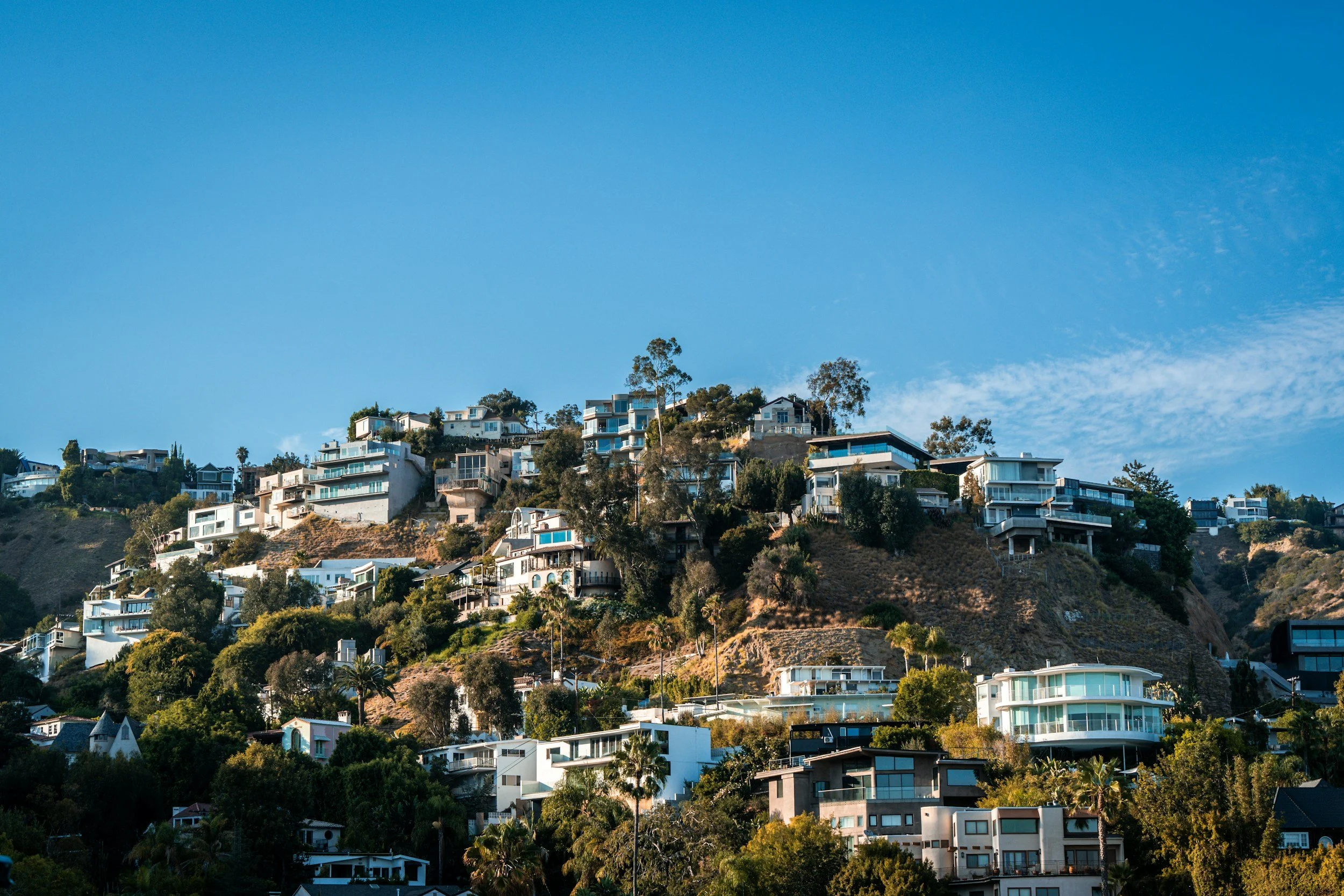 Hilly landscape with numerous modern white and dark-colored houses with large windows and terraces, surrounded by greenery and trees, under a clear blue sky.