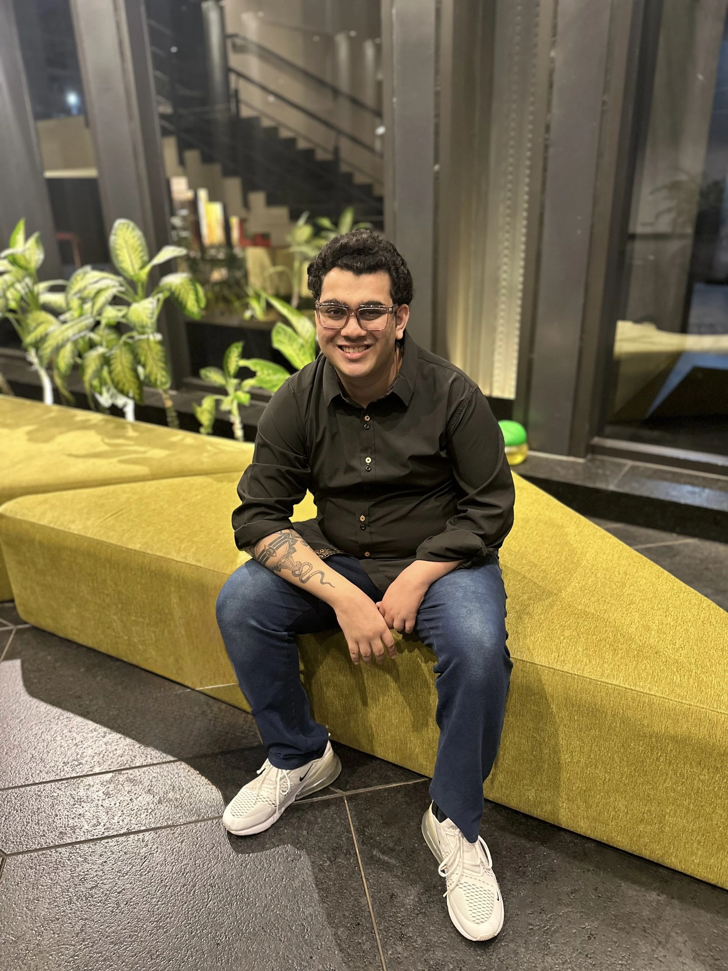 A young man with dark curly hair, glasses, and a tattooed arm, wearing a black shirt and jeans, sitting on a yellow upholstered bench in an indoor setting with plants and a metal staircase in the background.