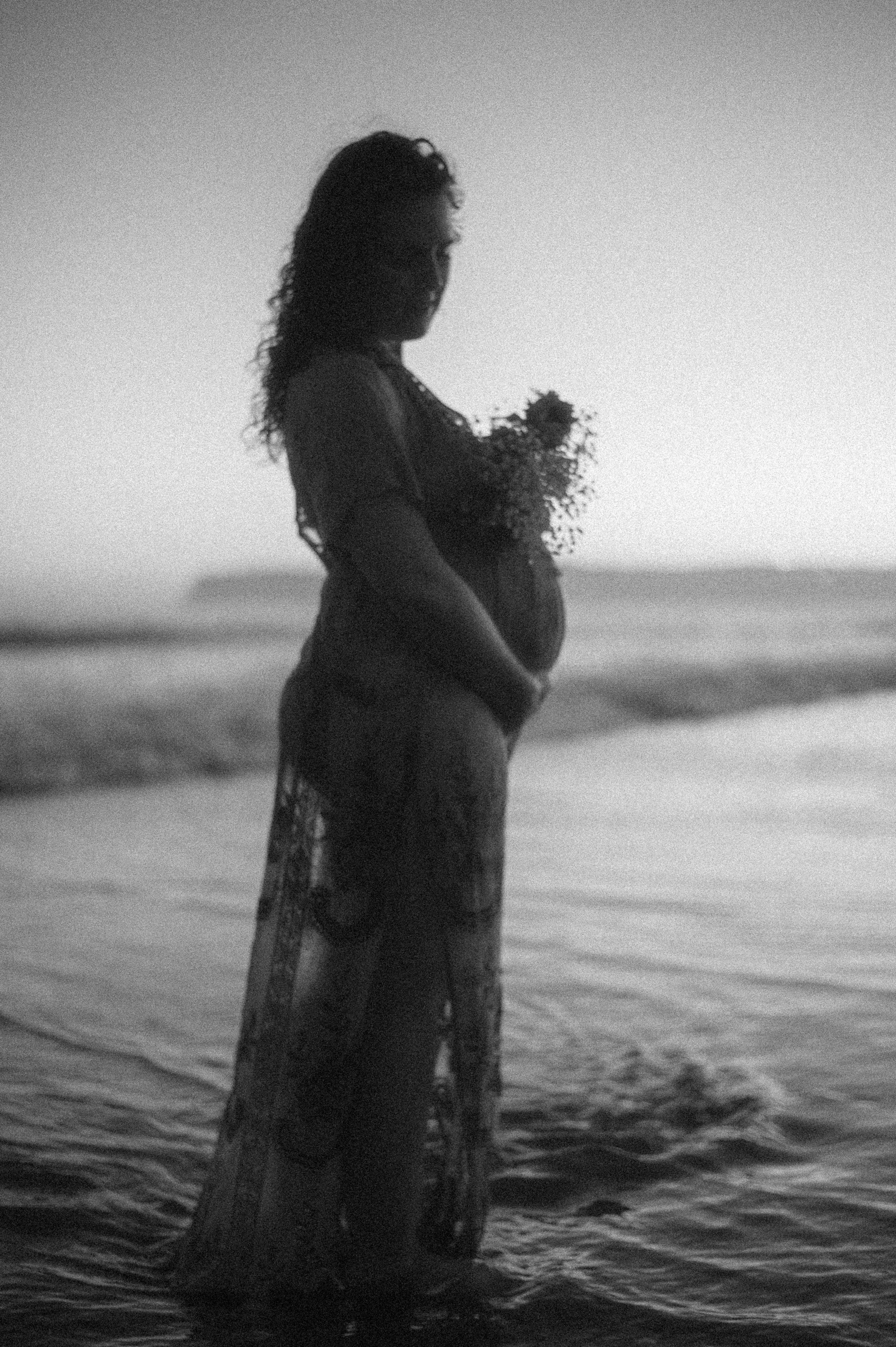 Silhouette of a pregnant woman standing on a beach at sunset with her hand in her pocket, holding a bouquet of flowers.