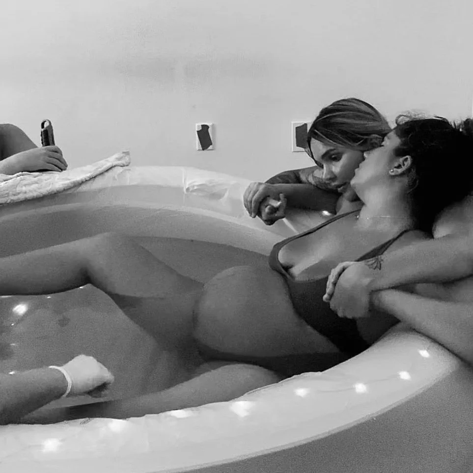 Two women lying in an inflatable hot tub, with one woman leaning over and touching the other's forehead, sharing an intimate moment.