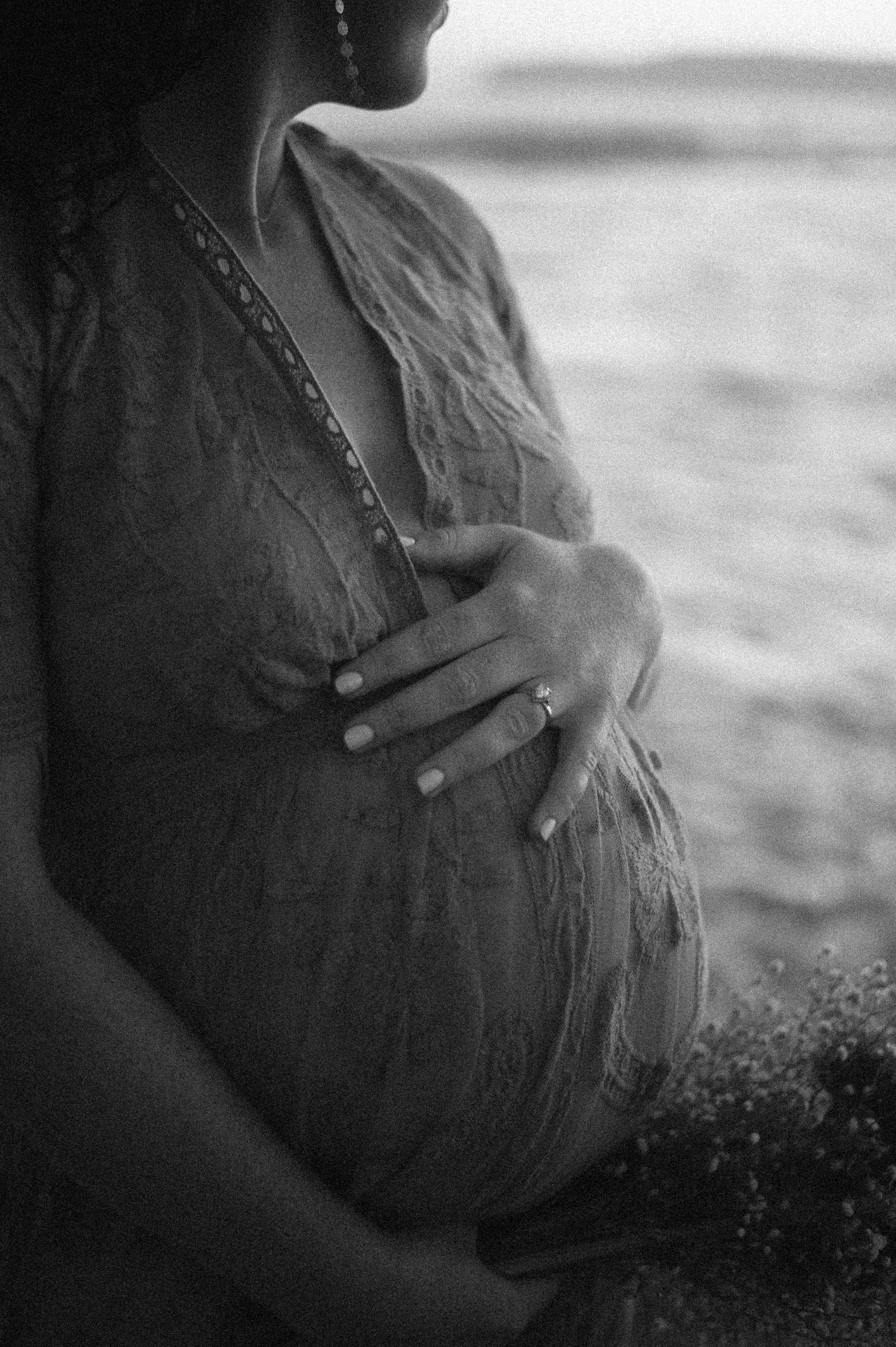 A pregnant woman in a lace top holding her belly near a body of water, with her face turned away from the camera.