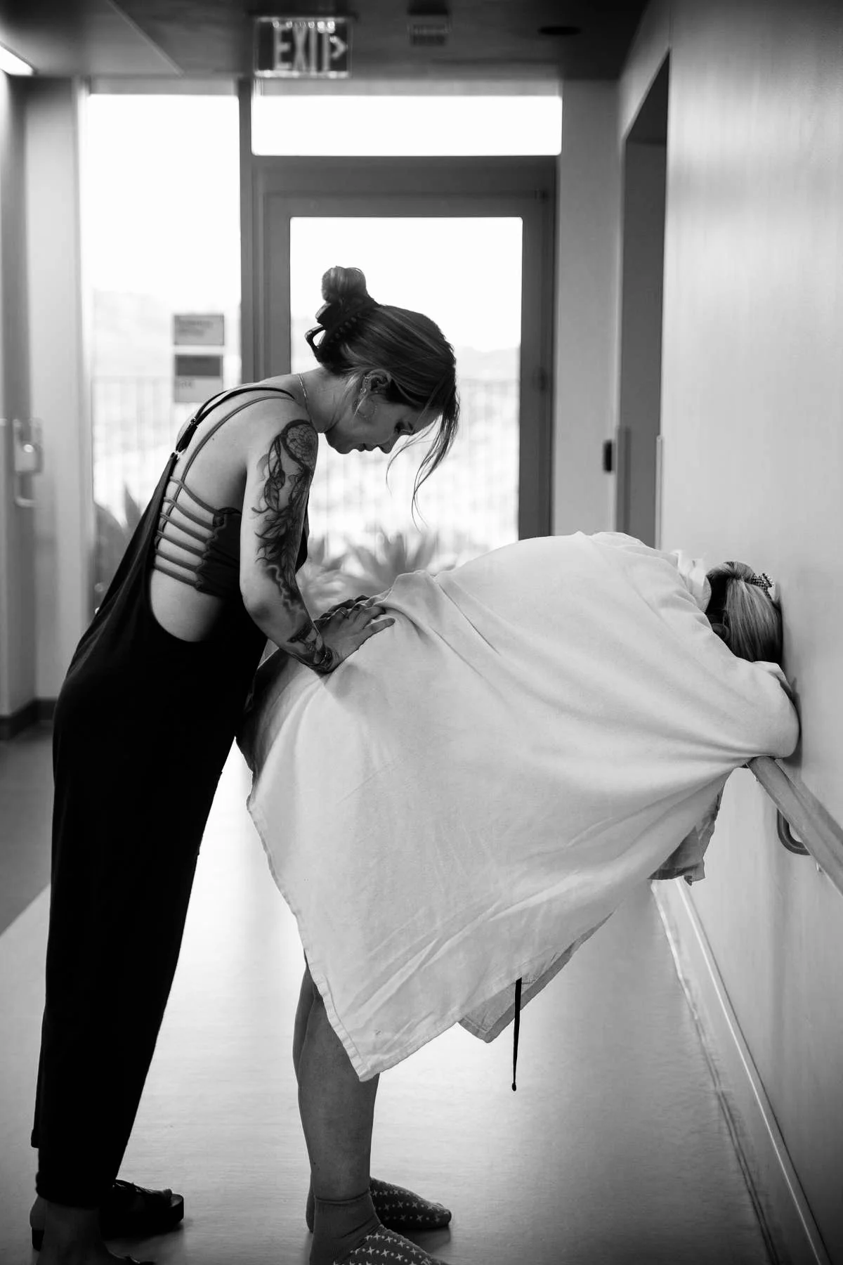A woman with tattoos on her arm leans over to comfort a woman lying in a hospital bed, who is resting her head on her crossed arms. The hospital room is lit with natural light coming through a glass door.