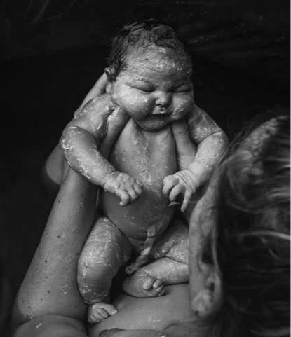 A baby being bathed, held by an adult's hands, with the adult partially visible and facing the baby.