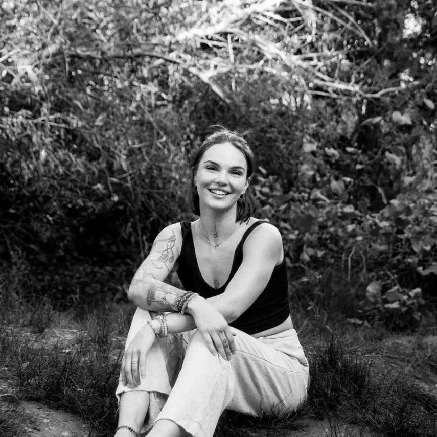 A young woman sitting outdoors in a natural setting with bushes and trees in the background. She is smiling, has tattoos on her arm, and is wearing a sleeveless top and light-colored pants.