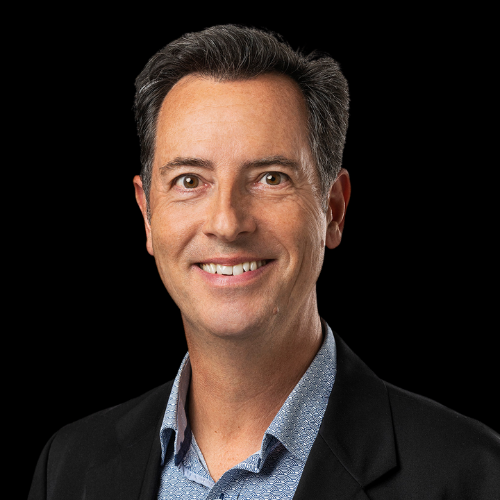 A professional headshot of a middle-aged man with short dark hair, wearing a dark suit jacket and a light blue shirt, smiling against a black background.