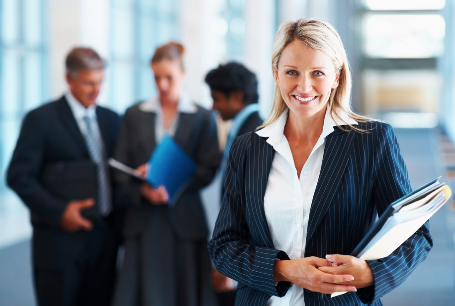 Businesswoman smiling and holding papers, with a group of professionals in the background in a modern office setting.