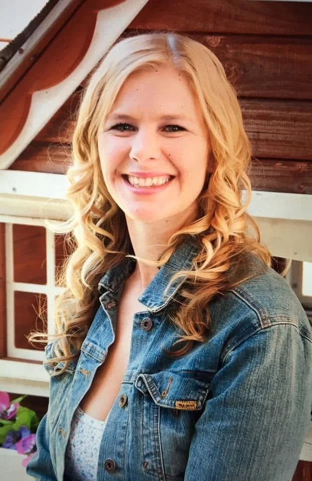 A young woman with long, curly blonde hair smiling at the camera, wearing a denim jacket, outdoors with a wooden structure and window in the background.
