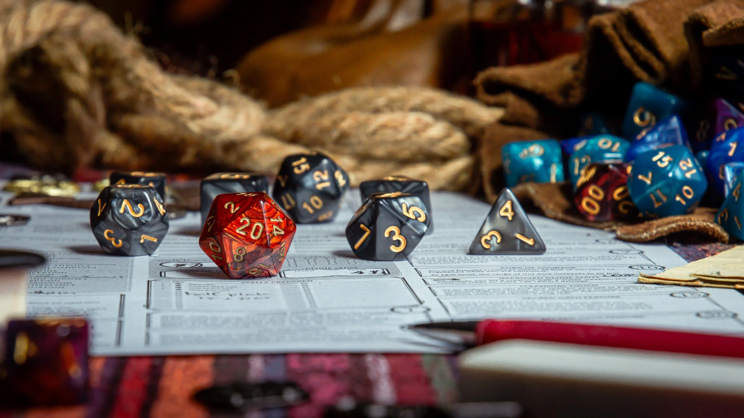 A tabletop with various gaming dice, including a red 20-sided die, blue 20-sided dice, black 20-sided dice, and a white 4-sided die, placed on top of a game sheet surrounded by papers, a pen, and background items like ropes and bark.
