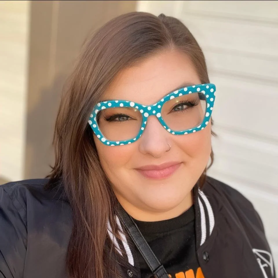 A woman with long brown hair wearing blue polka dot glasses, a black jacket, and a black shirt with orange and yellow text, smiling at the camera.
