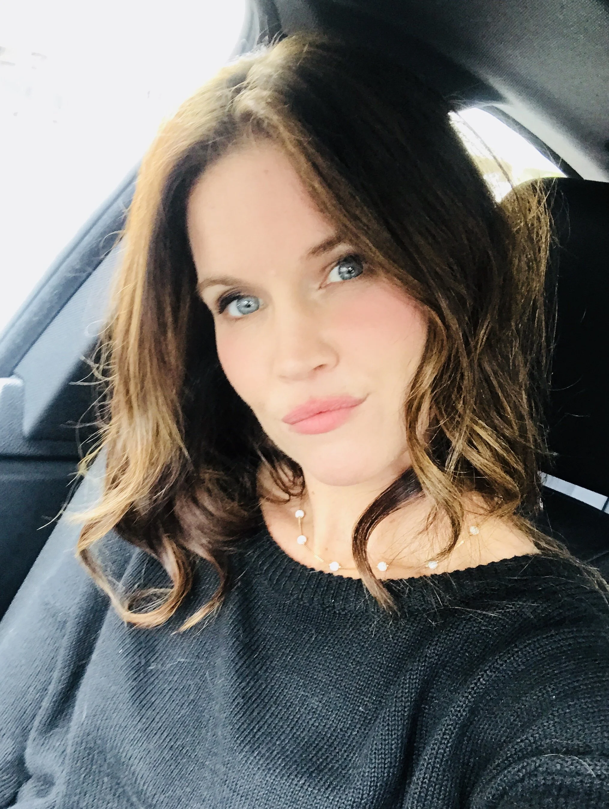 A woman with wavy brown hair and blue eyes sitting in a car. She is wearing a black top and a pearl necklace. The photo is a close-up selfie taken inside a vehicle.