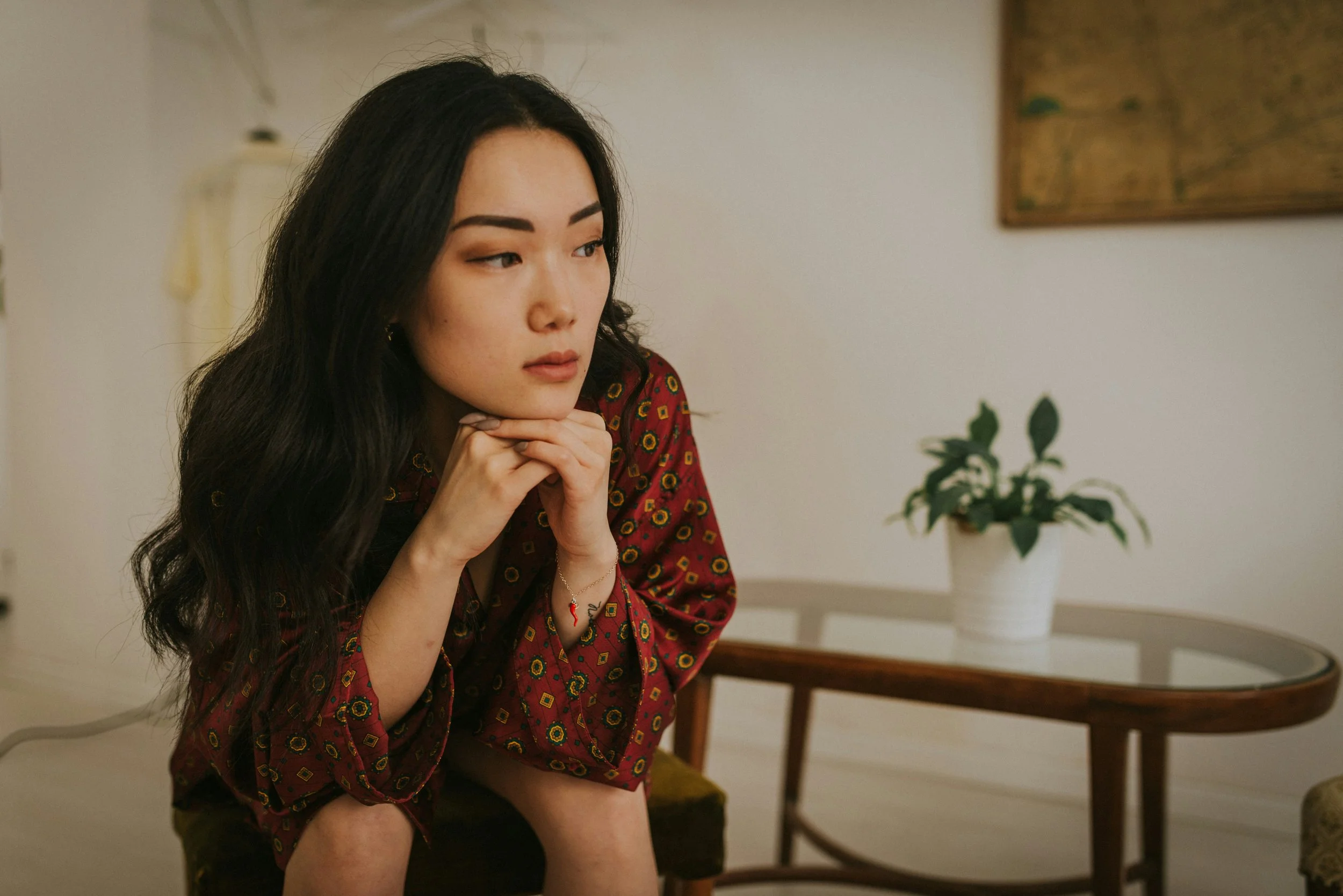 A woman with long dark hair sitting on a chair, resting her chin on her hands, looking thoughtfully to the side. She is wearing a red patterned top, and there is a side table with a potted plant in the background.