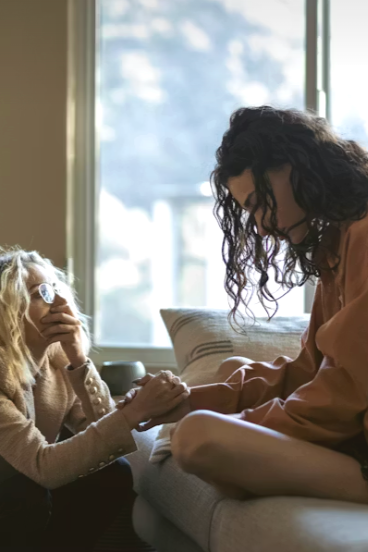 Two women sitting on a bed, holding hands, and having a heartfelt conversation in a sunlit room.