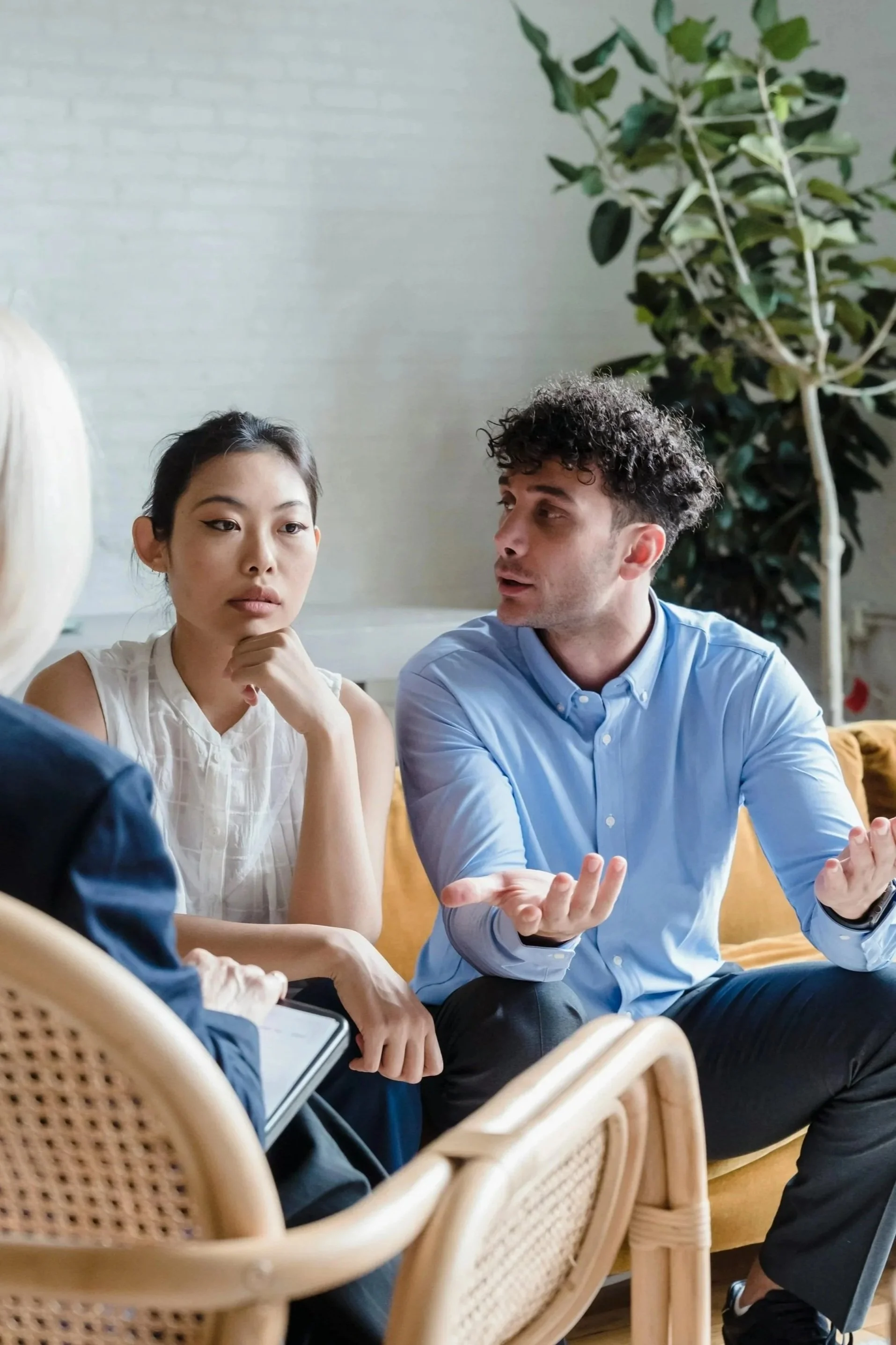 Group of three people engaged in a discussion in an office or waiting area, with chairs and a plant in the background.