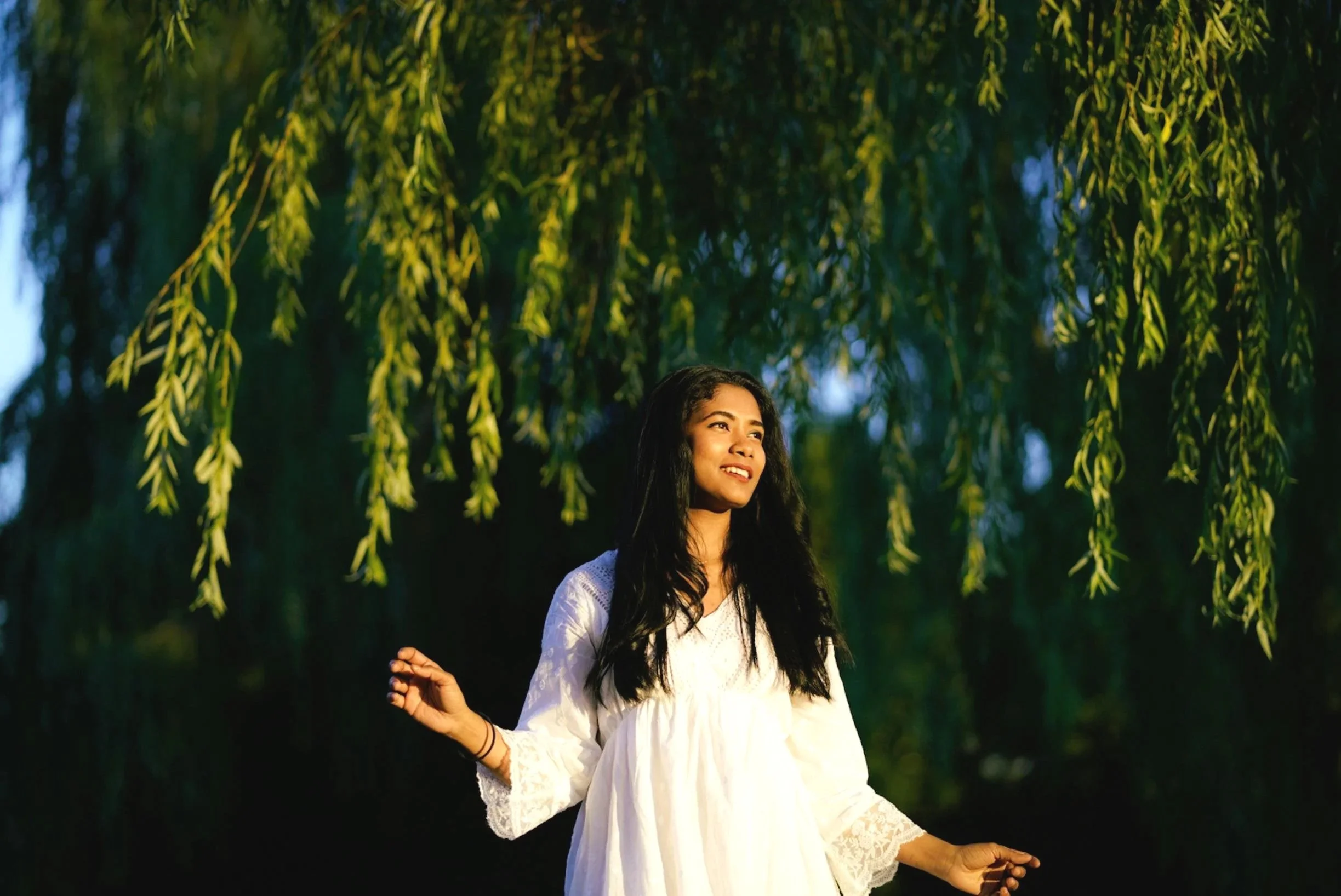 A young woman with long dark hair wearing a white dress standing outdoors under green hanging tree branches, looking to the side with a smile.