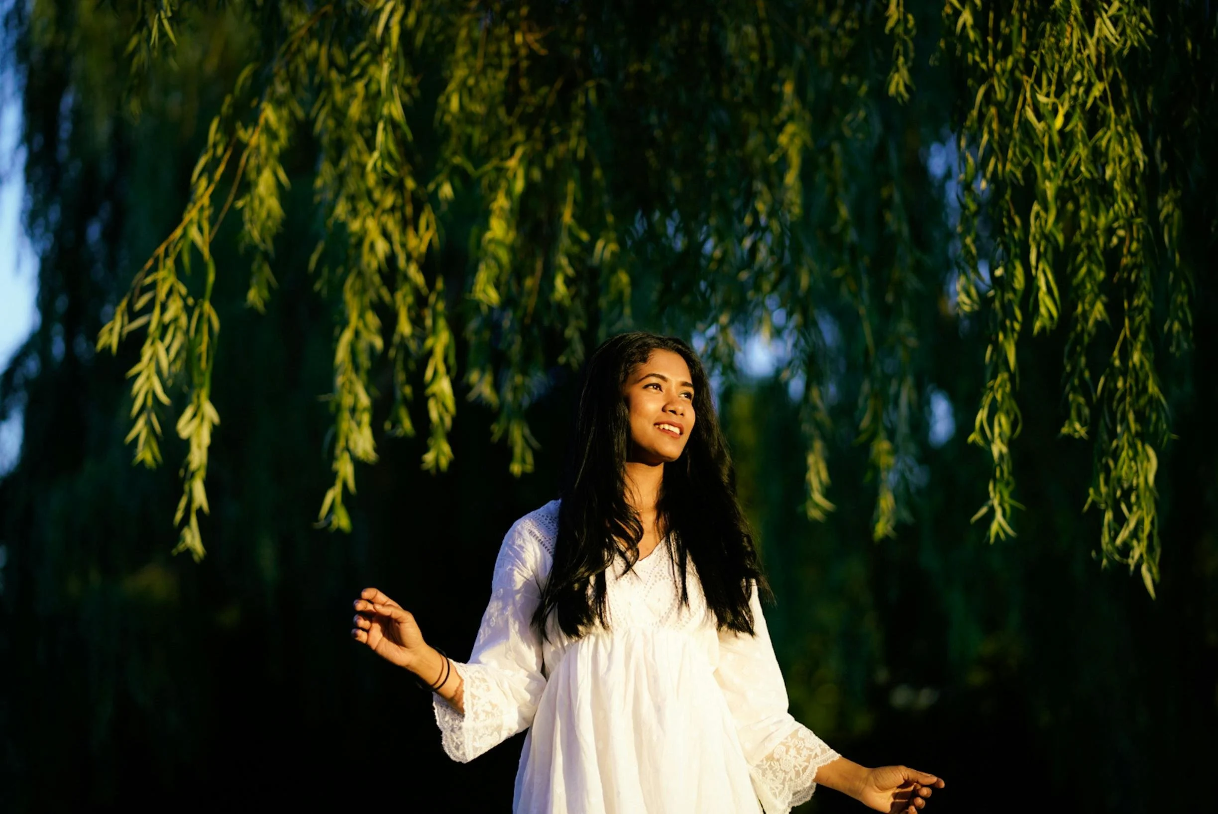 A woman in a white dress standing outdoors beneath a large hanging tree with green leaves, smiling as the sunlight filters through.