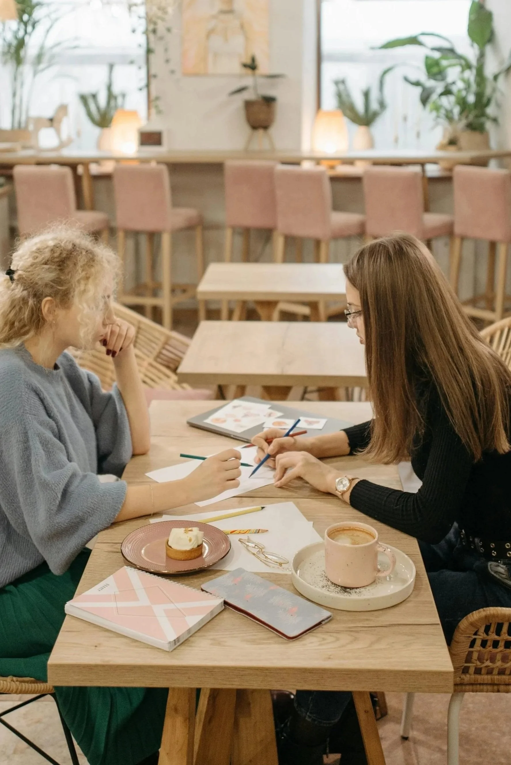 Two women sitting at a wooden table in a cozy cafe, discussing and working on documents with notebooks, pens, and a tablet. One has curly blonde hair and the other has straight brown hair. There are coffee cups and a small cake on the table.
