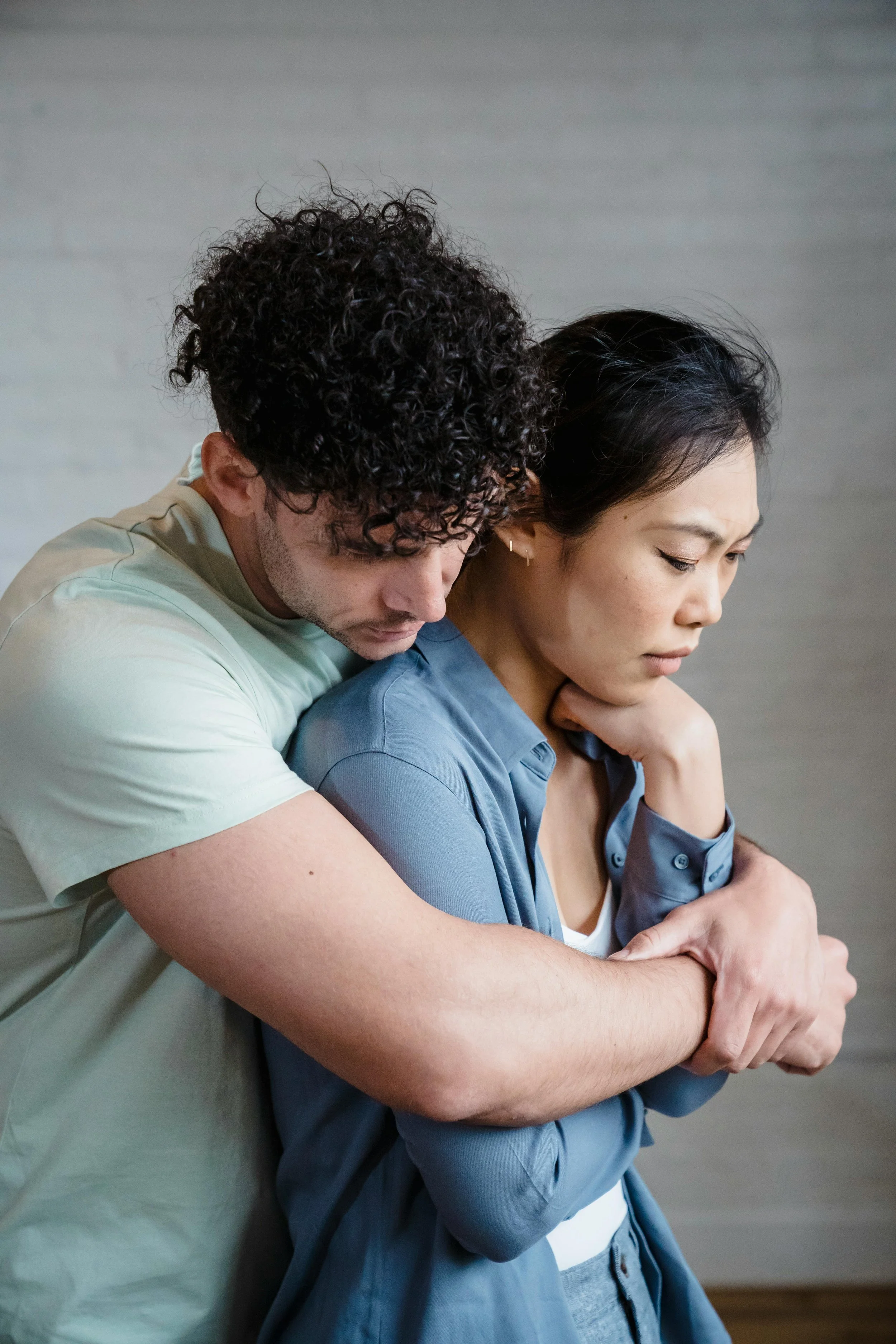 A man gently comforting a woman who looks distressed, with his arms wrapped around her shoulders from behind.