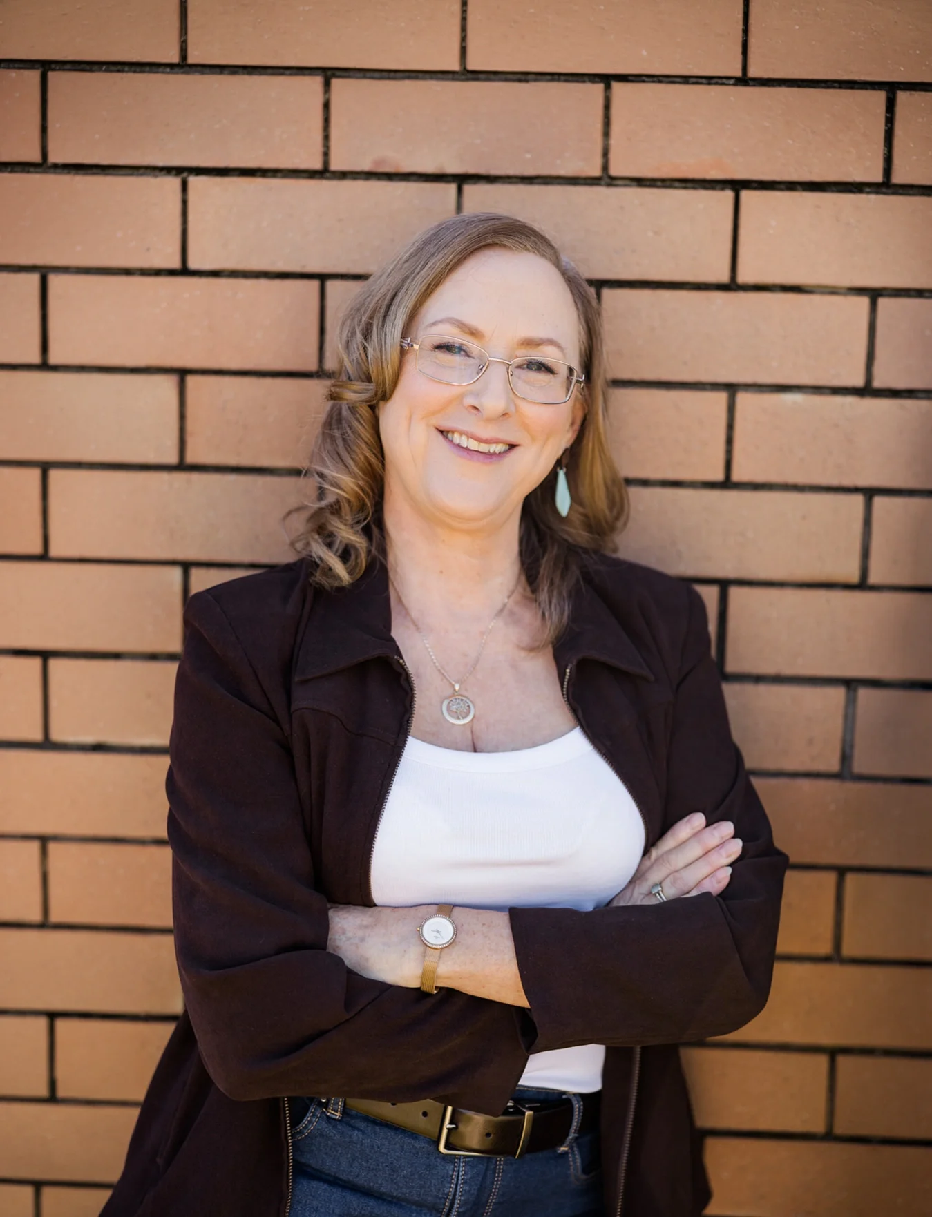 A woman with shoulder-length wavy hair, wearing glasses, smiling, standing with her arms crossed in front of a brick wall.