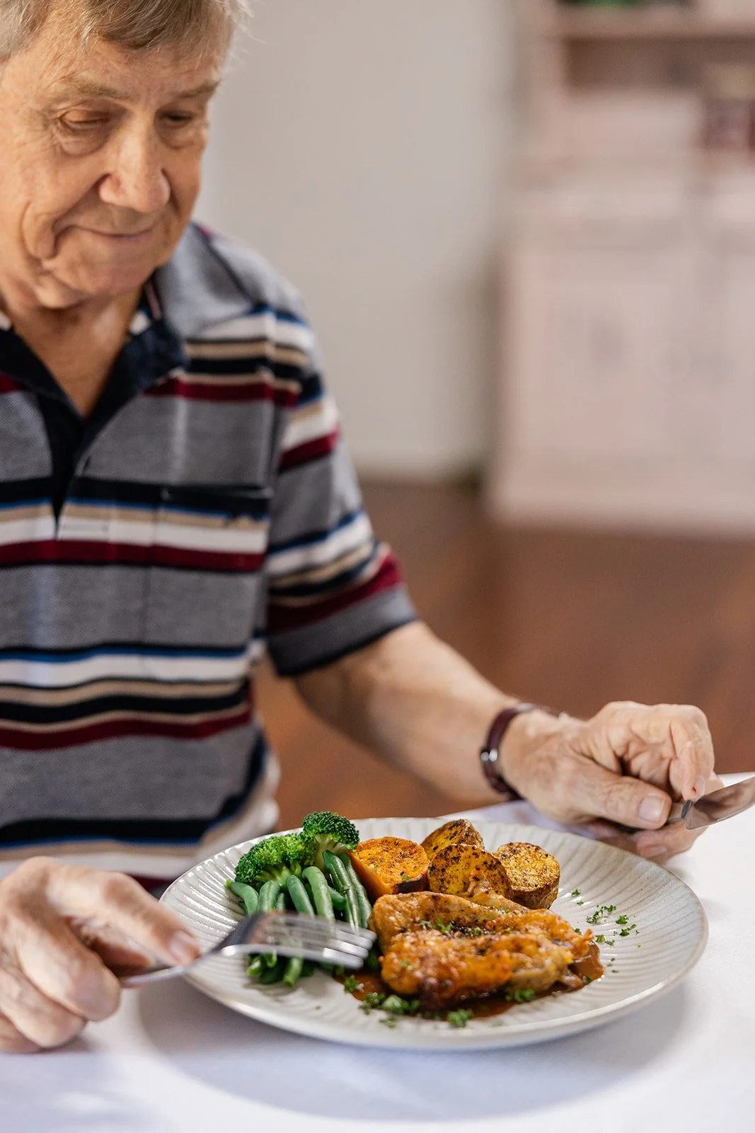 Older man with striped shirt sitting at a table with a plate of food, including chicken, sweet potatoes, broccoli, and green beans.