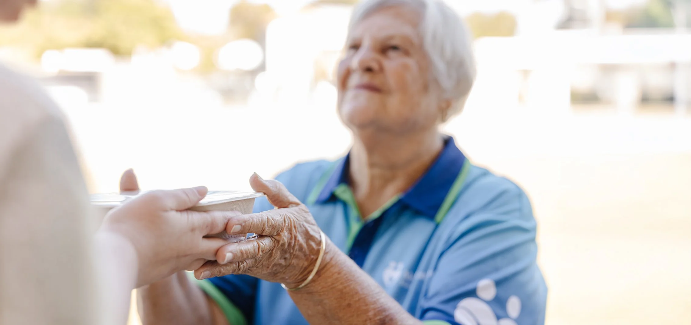 An elderly woman with short gray hair wearing a blue shirt handing a bowl of food to someone off-camera in an outdoor setting with sunlight and blurred background.