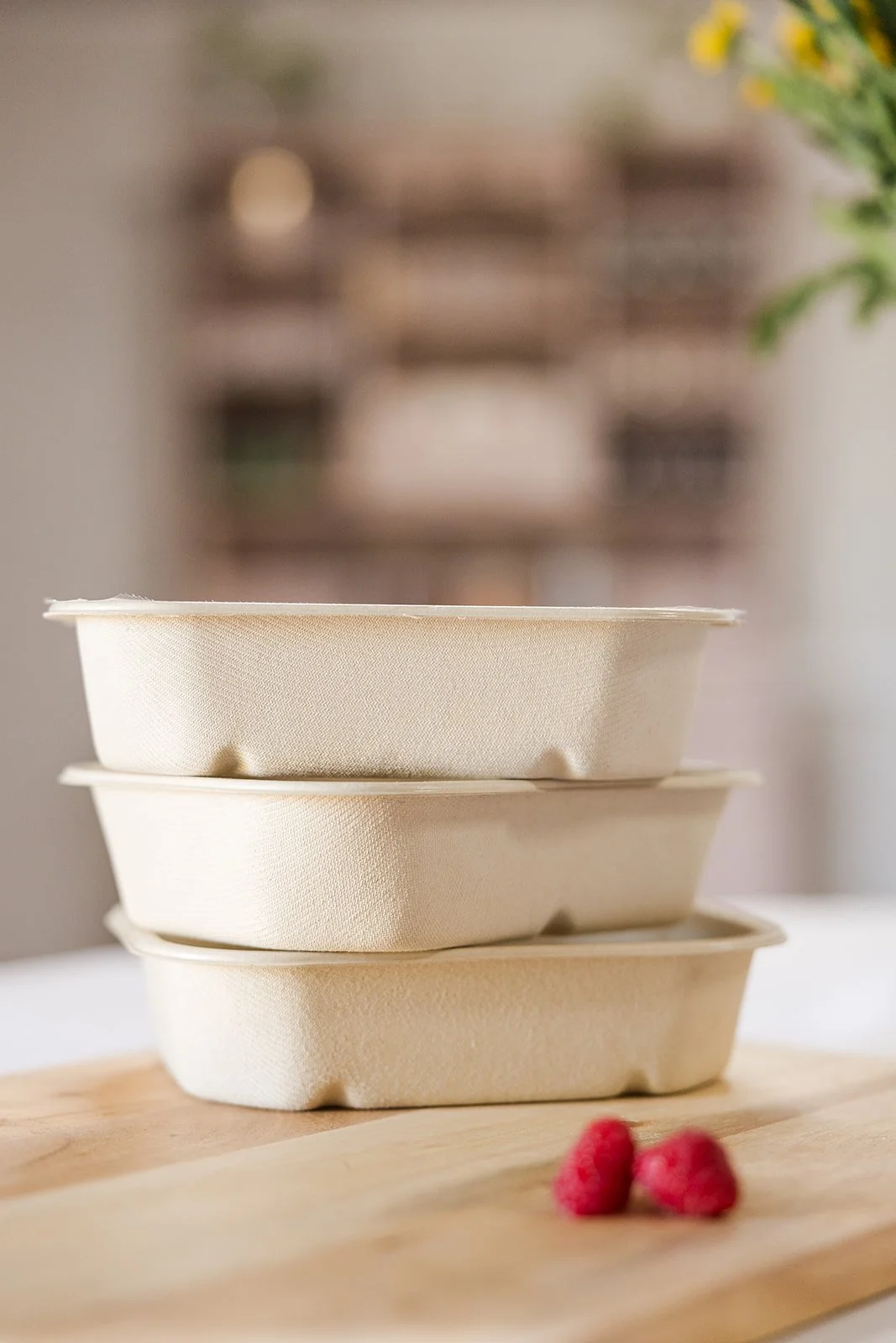 Three stacked beige-colored food containers on a wooden surface with two raspberries in the foreground and a blurred background of a shelf and some greenery.