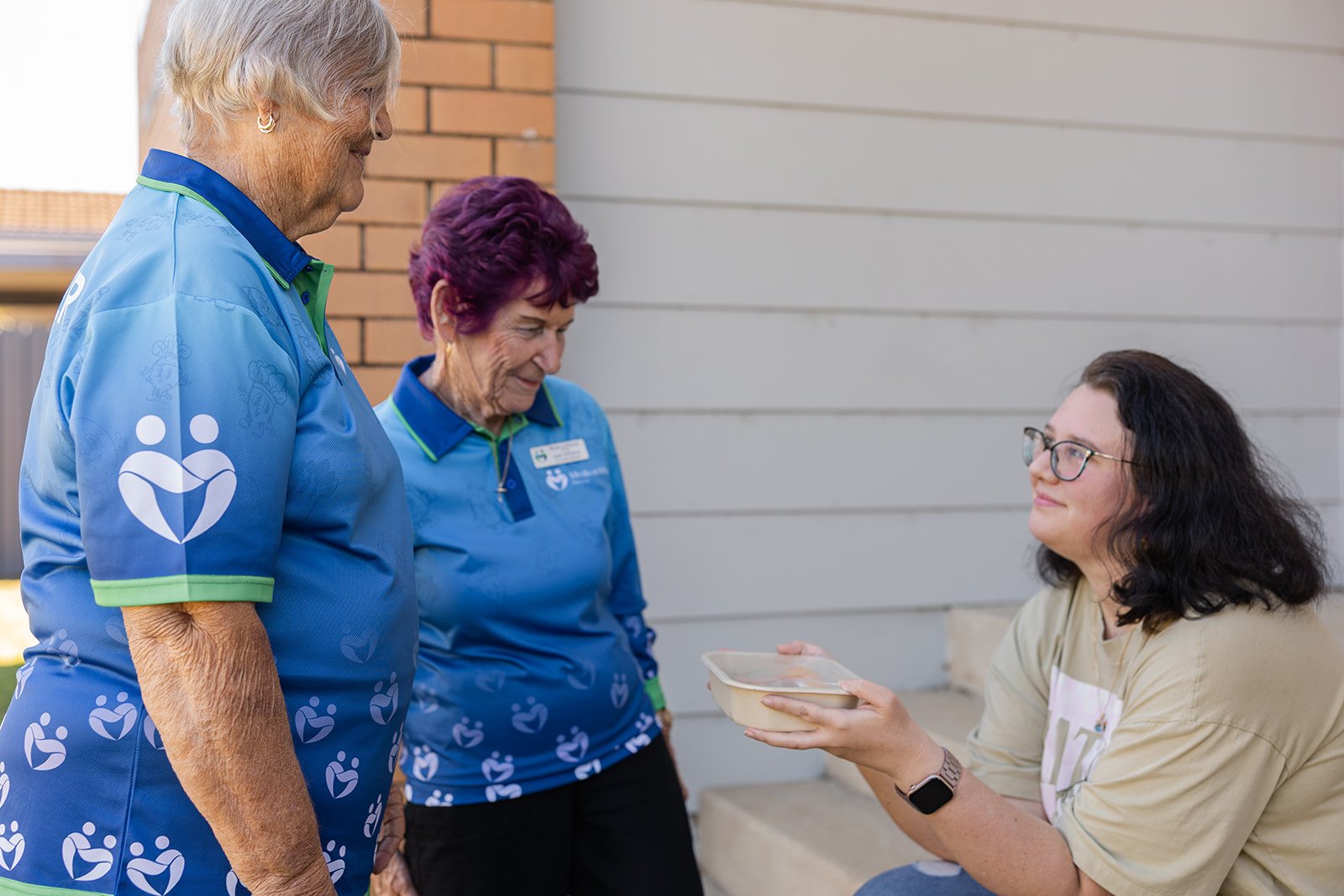 Four women, two standing and two sitting, engaging in a conversation outdoors. One woman is handing a container of food to the seated woman with dark curly hair and glasses. The women standing are wearing matching blue shirts with a logo, while the seated woman is wearing a beige shirt.