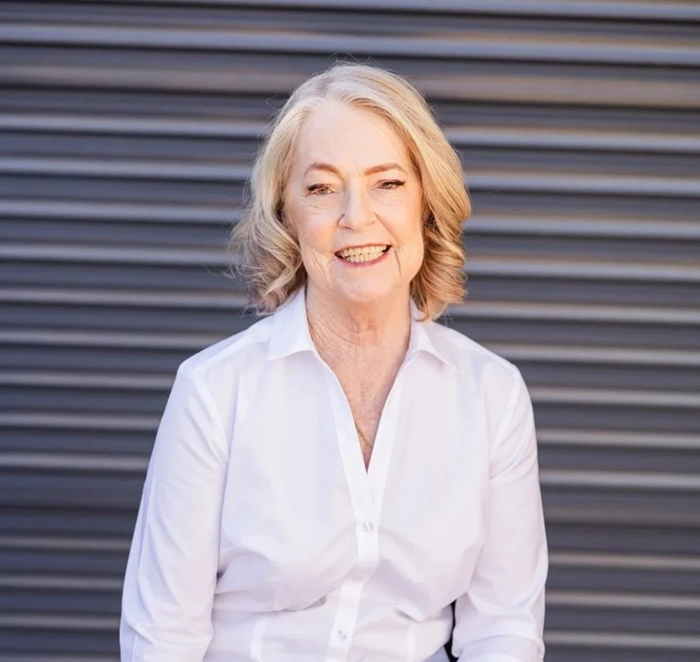 A middle-aged woman with blonde hair is smiling in front of a metallic, horizontal-paneled background. She is wearing a white button-up shirt.