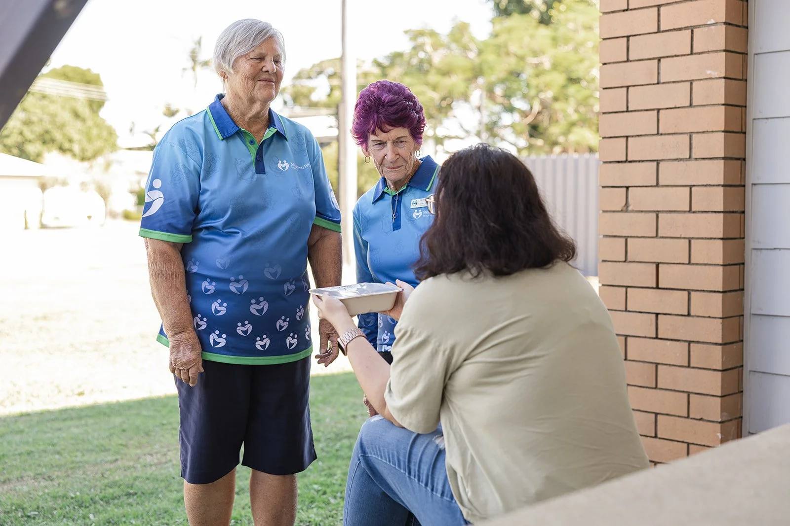 Two elderly women in matching light blue shirts with logos are standing outside on the grass, looking at a woman sitting on a step holding a food container. There's a brick wall and a wooden house siding visible on the right.