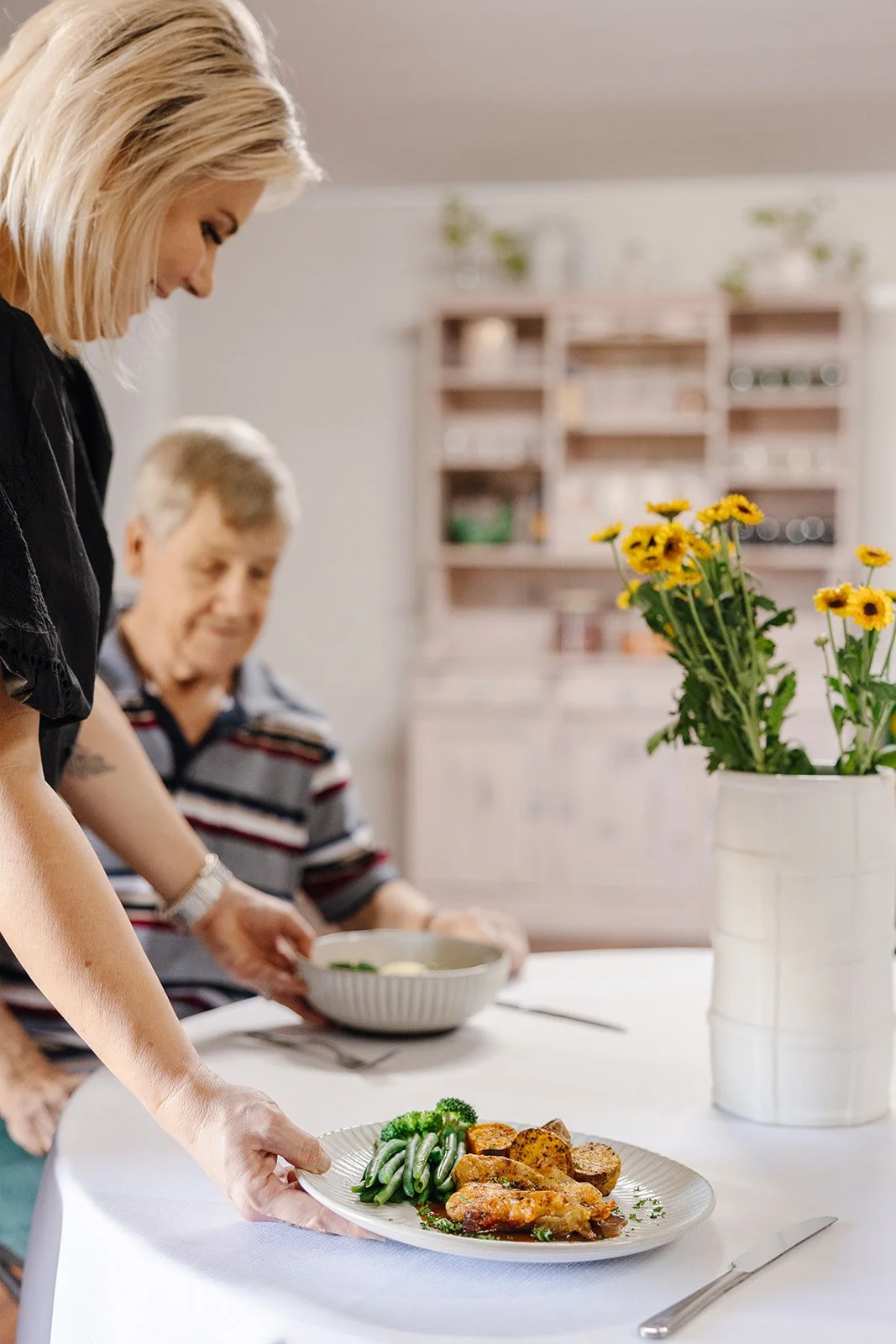 A woman is setting a plate of food that includes green beans, roasted potatoes, and chicken on a dining table. An older man is in the background holding a bowl. A white vase with yellow flowers is on the table.