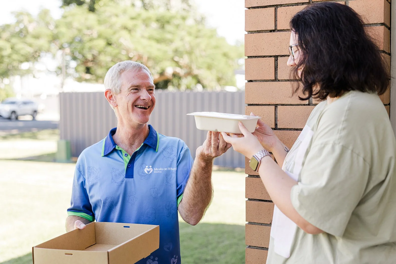 A smiling man in a blue volunteer shirt hands a food container to a woman standing outside next to a brick wall. The man is holding a cardboard box, and both are engaging in a friendly exchange.