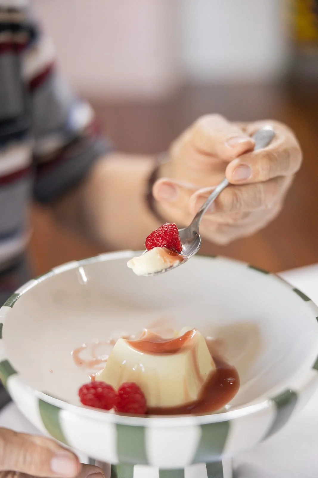 Close-up of an elderly person's hand holding a spoon with a raspberry and a dollop of cream, about to eat a serving of pudding topped with caramel sauce and raspberries in a striped bowl.