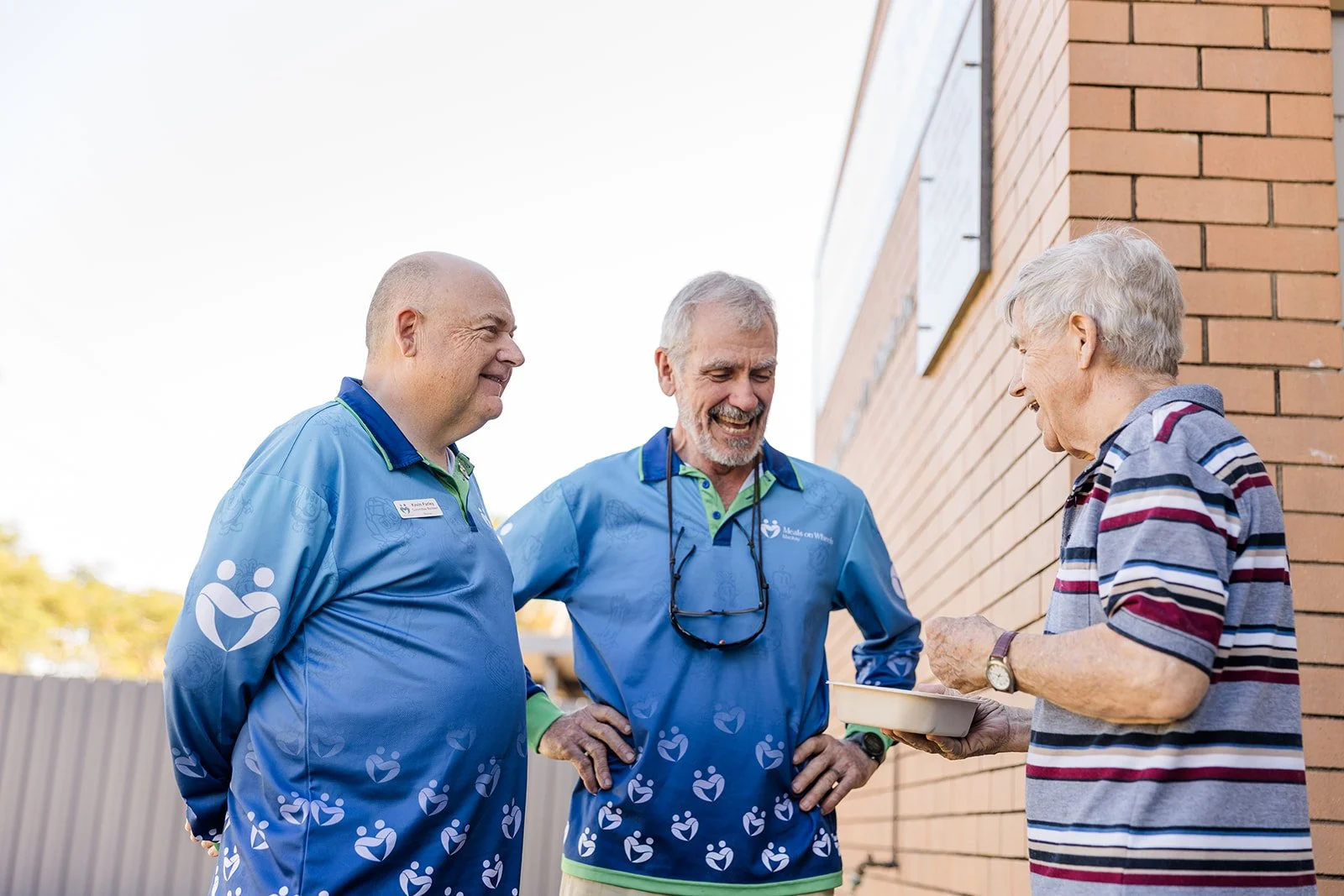 Two men smiling and talking with an elderly man who is holding a tray of food outdoors near a brick wall.