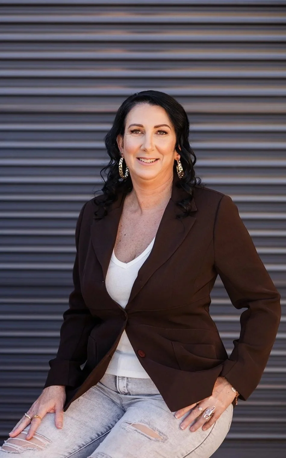 A woman with black, curly hair, wearing a brown blazer, white top, ripped jeans, and large earrings, posing in front of a metallic, ribbed background.