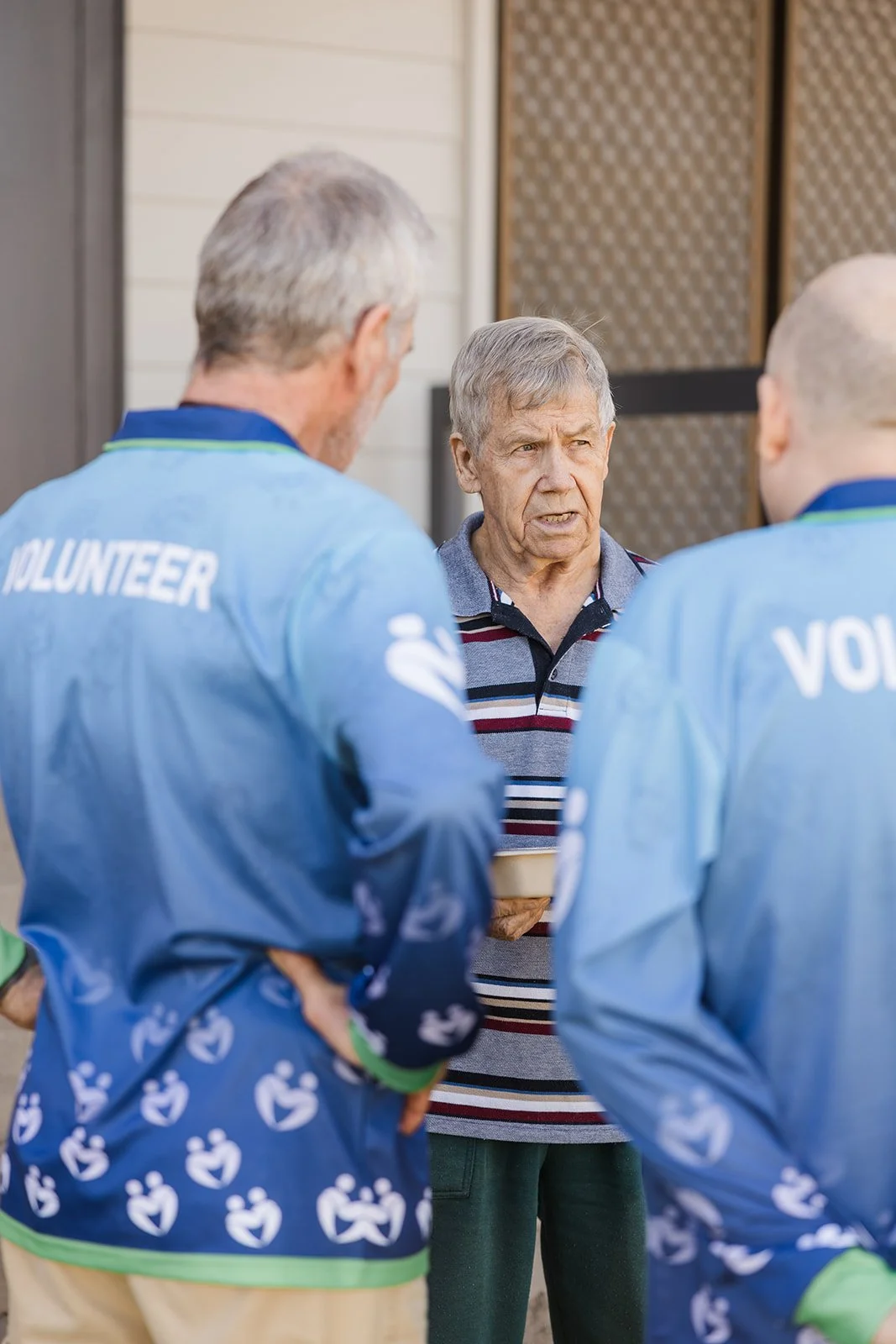 An elderly man in a striped polo shirt speaking to two volunteers in light blue uniforms with 'VOLUNTEER' printed on the back, outside a house.