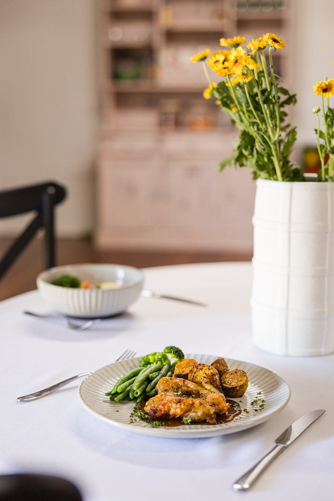 A dinner plate with chicken, green beans, broccoli, and roasted potatoes on a white tablecloth, with a fork and knife on either side, and a white vase with yellow flowers in the background.