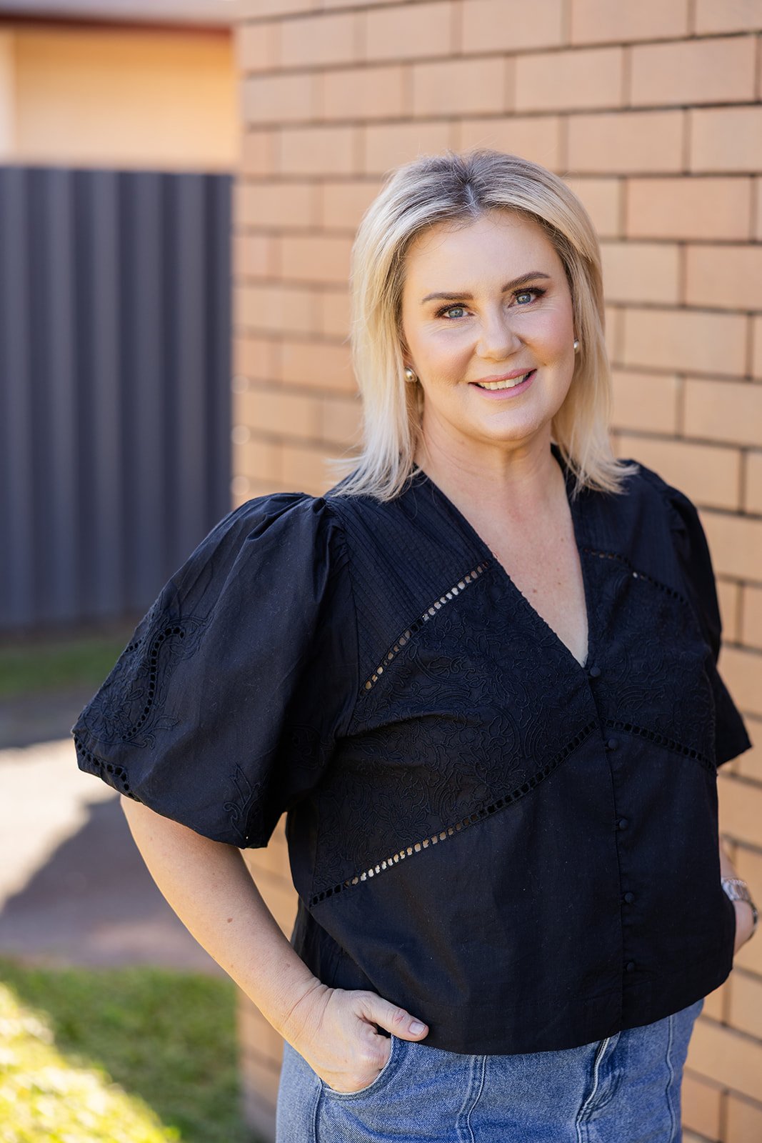 A blonde woman with blue eyes, wearing a black blouse and blue jeans, standing outdoors in front of a brick wall, smiling at the camera.