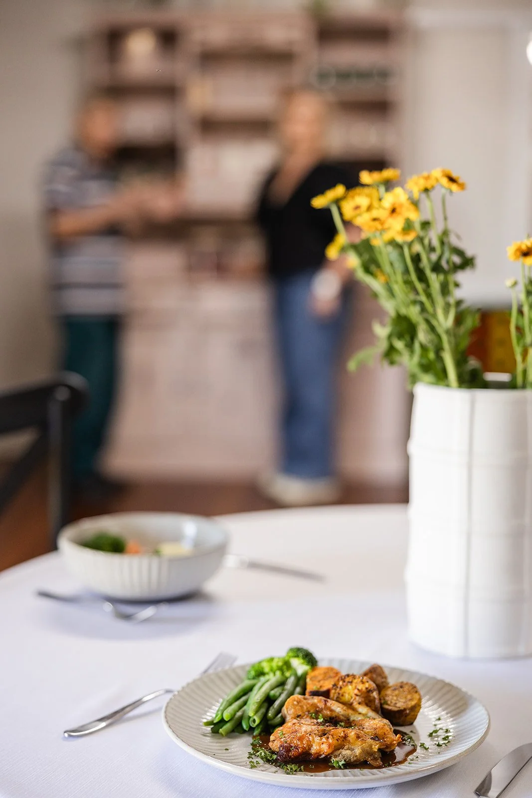 A dinner plate with cooked chicken, green beans, and potatoes on a white tablecloth, with a bowl of salad and a vase of yellow flowers in the background.