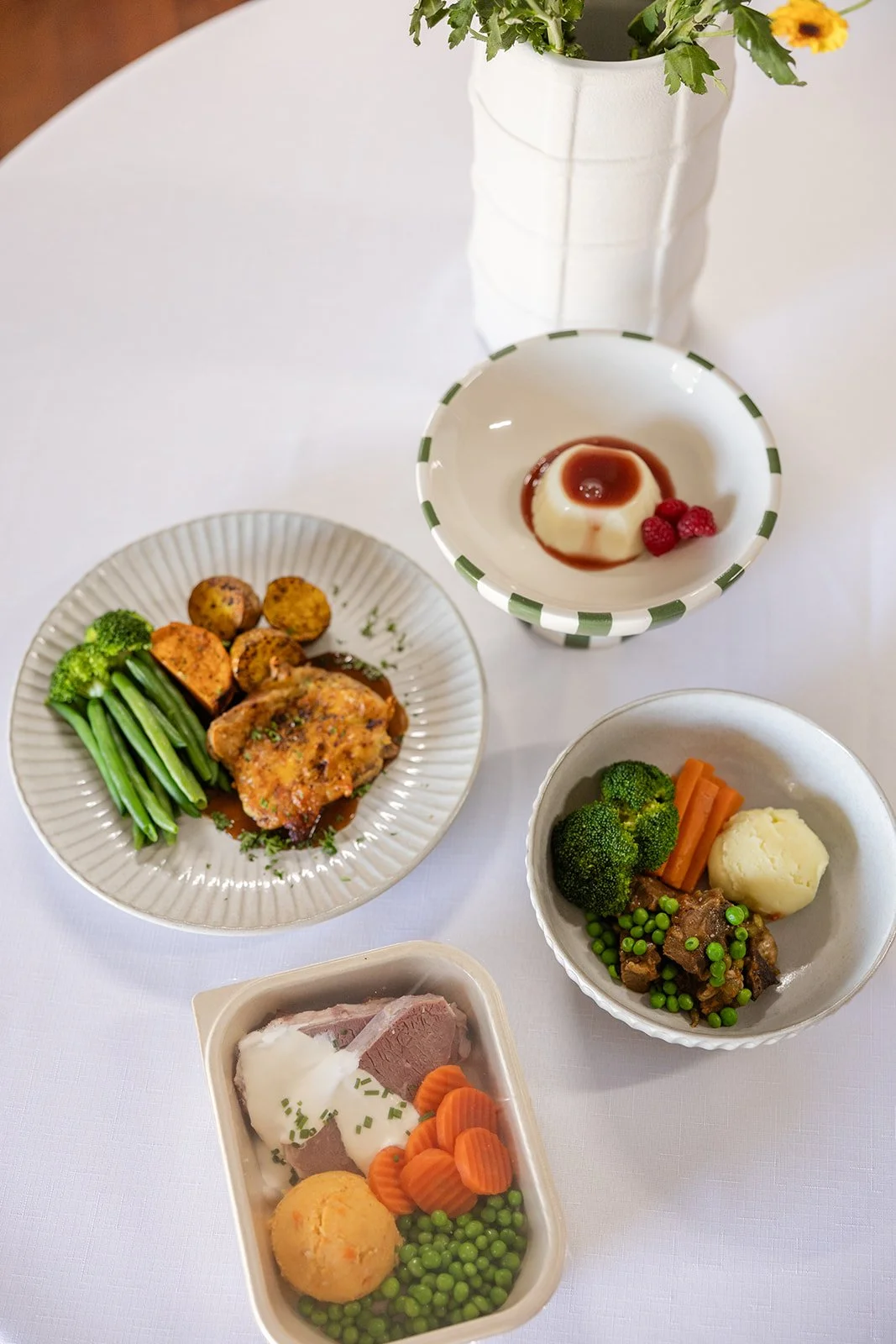 A table with various dishes including a plate of cooked vegetables, a bowl with pudding and raspberries, a dish with broccoli, carrots, mashed potatoes, and beef stew, and a container with sliced meat, carrots, green peas, a meatball, and gravy, with a white vase holding flowers in the background.