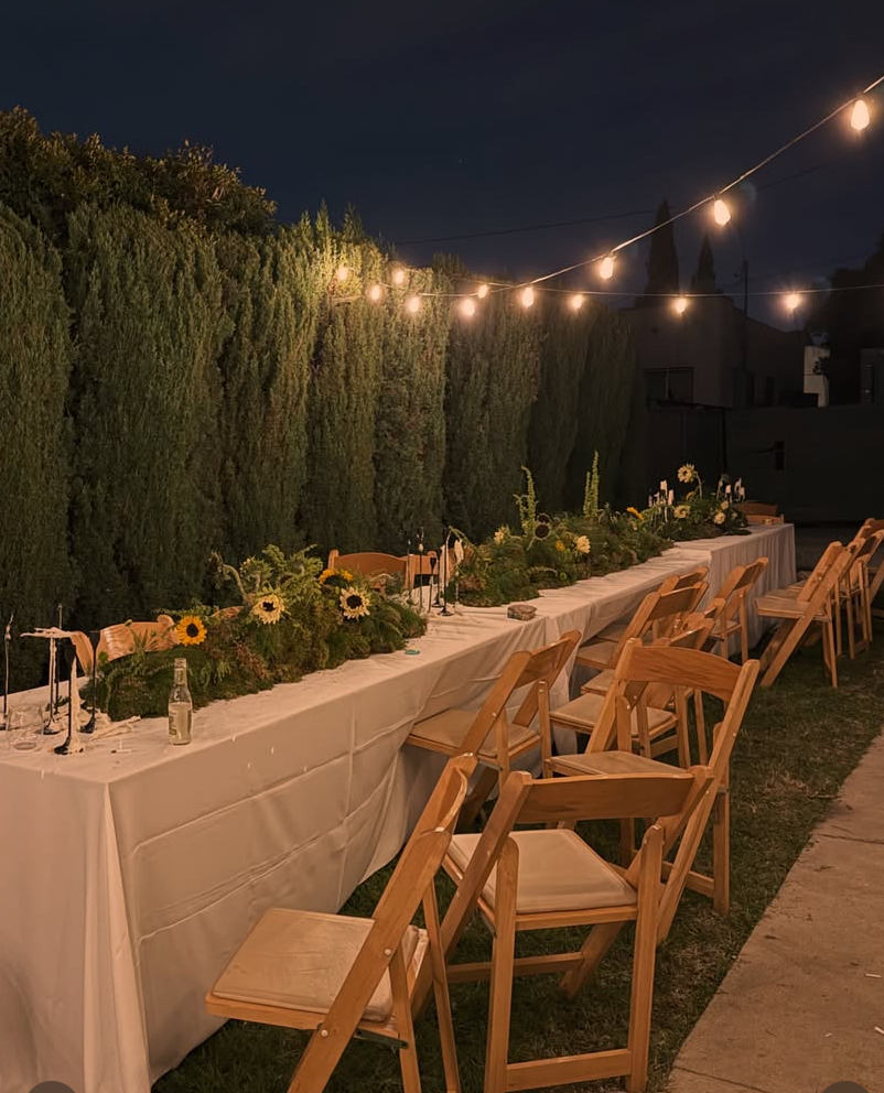 Outdoor evening dining setup with a long table covered with a white tablecloth, decorated with flower arrangements, sunflowers, and greenery, illuminated by string lights, wooden chairs around the table, and tall cypress trees in the background.