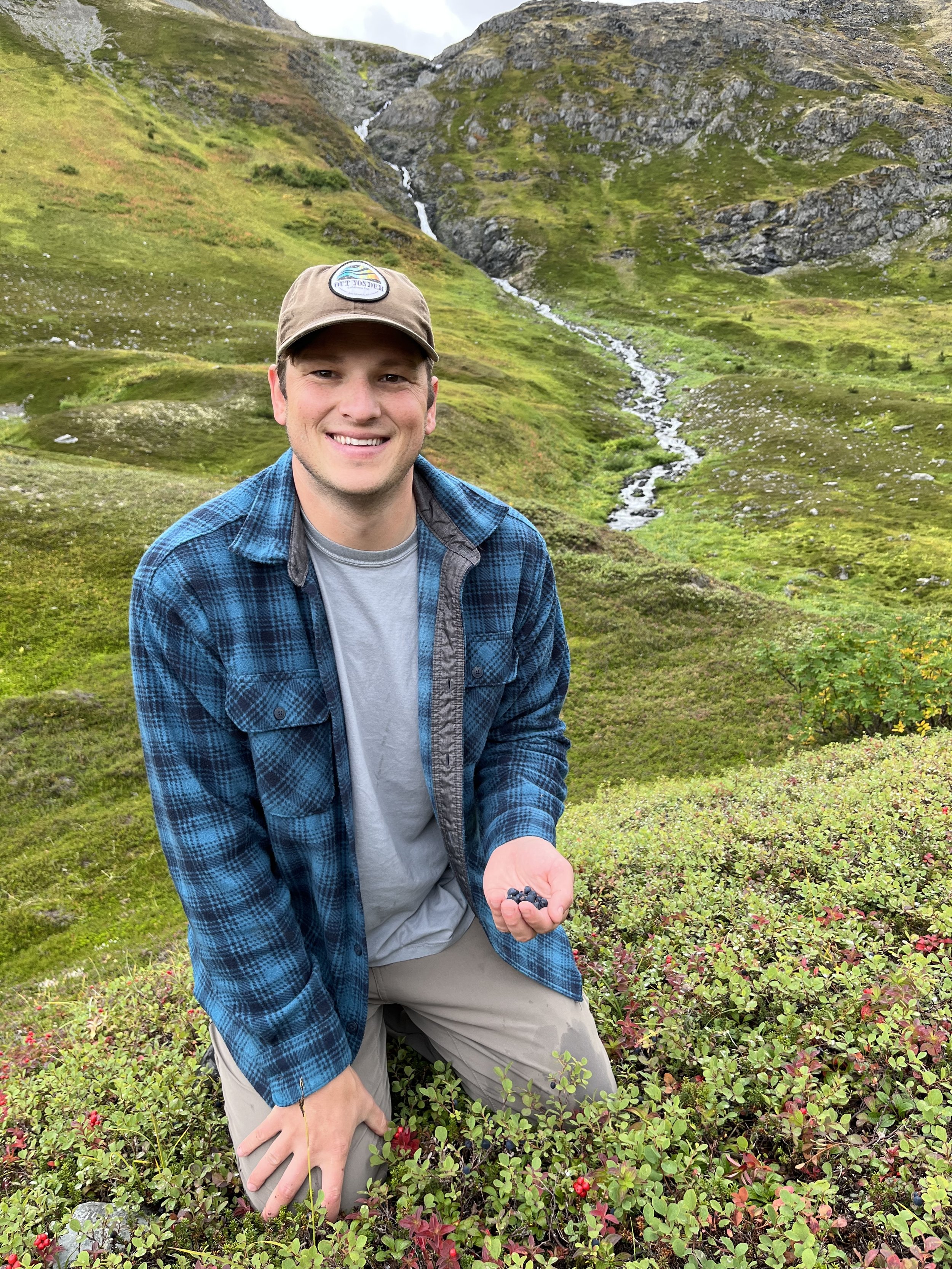 A young man in outdoor attire holding blueberries in his right hand, kneeling in a lush green field with low shrubs and blueberries, mountains in the background with a small stream flowing down the hillside.