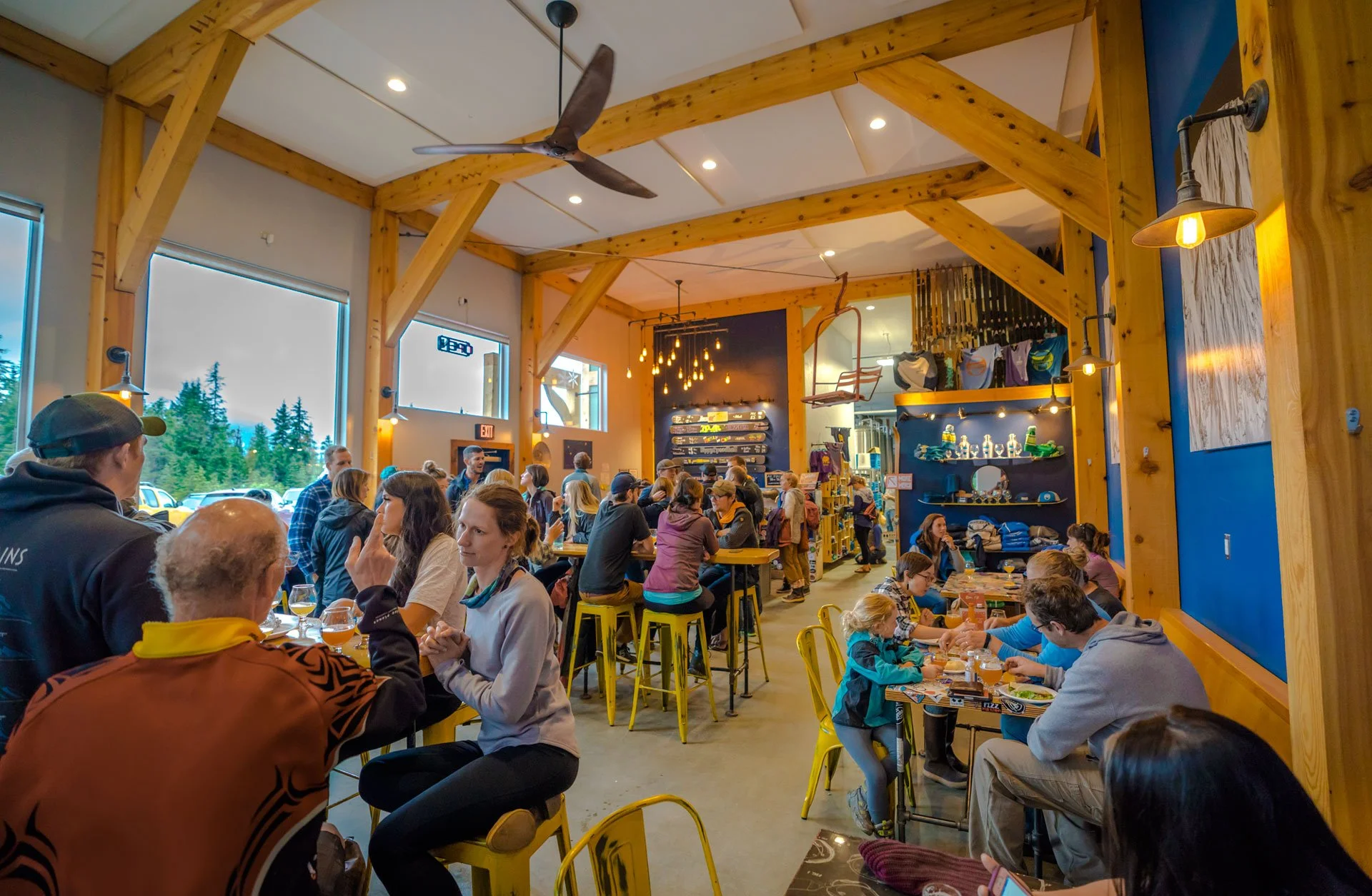 People dining inside a well-lit wooden restaurant with large windows and a high ceiling, decorated with industrial-style wall lights and hanging Edison bulbs.