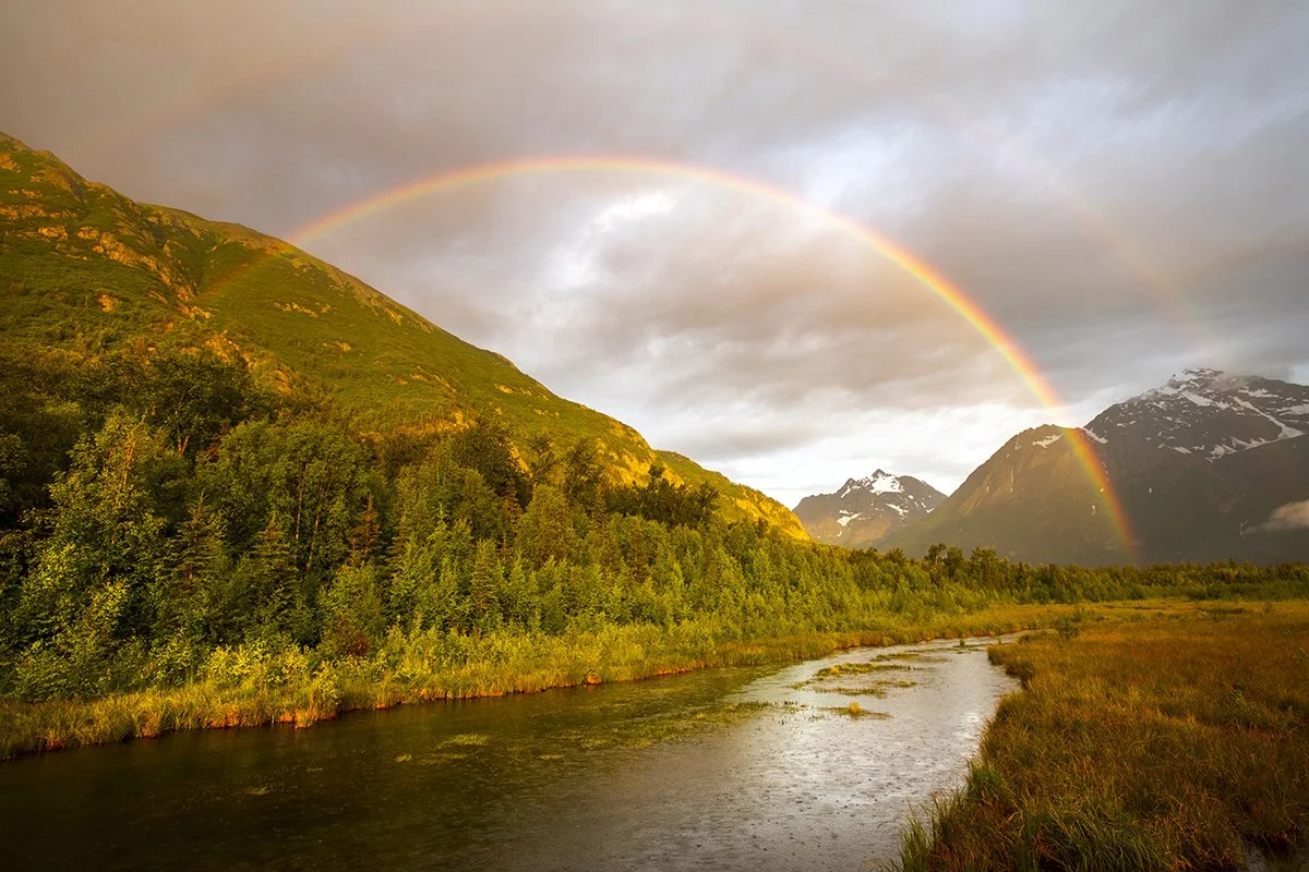 Landscape with a river flowing through a grassy field, surrounded by green mountains with snow-capped peaks, and a double rainbow in the cloudy sky.