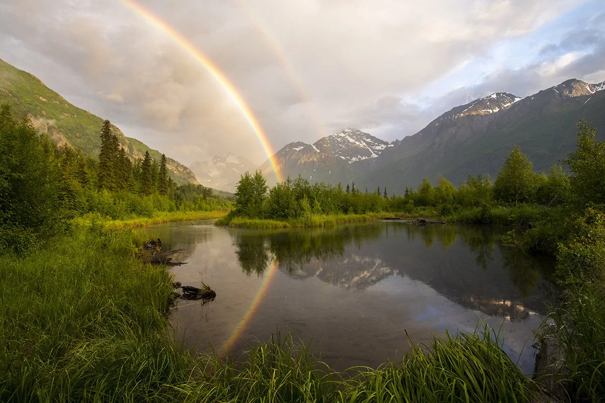 A scenic landscape featuring a rainbow over a mountain river surrounded by lush green trees and snow-capped mountains in the background.