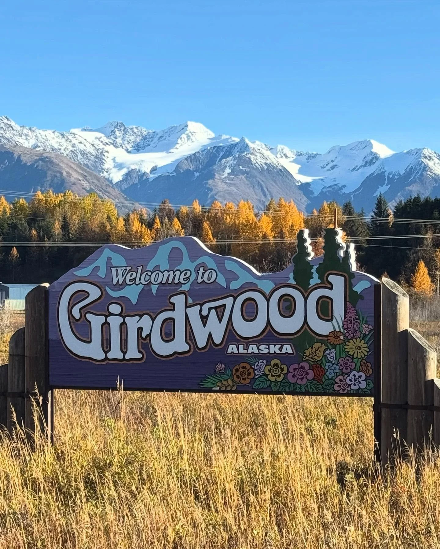 A colorful welcome sign for Girdwood, Alaska, with a backdrop of snow-capped mountains, trees with fall foliage, and a clear blue sky.